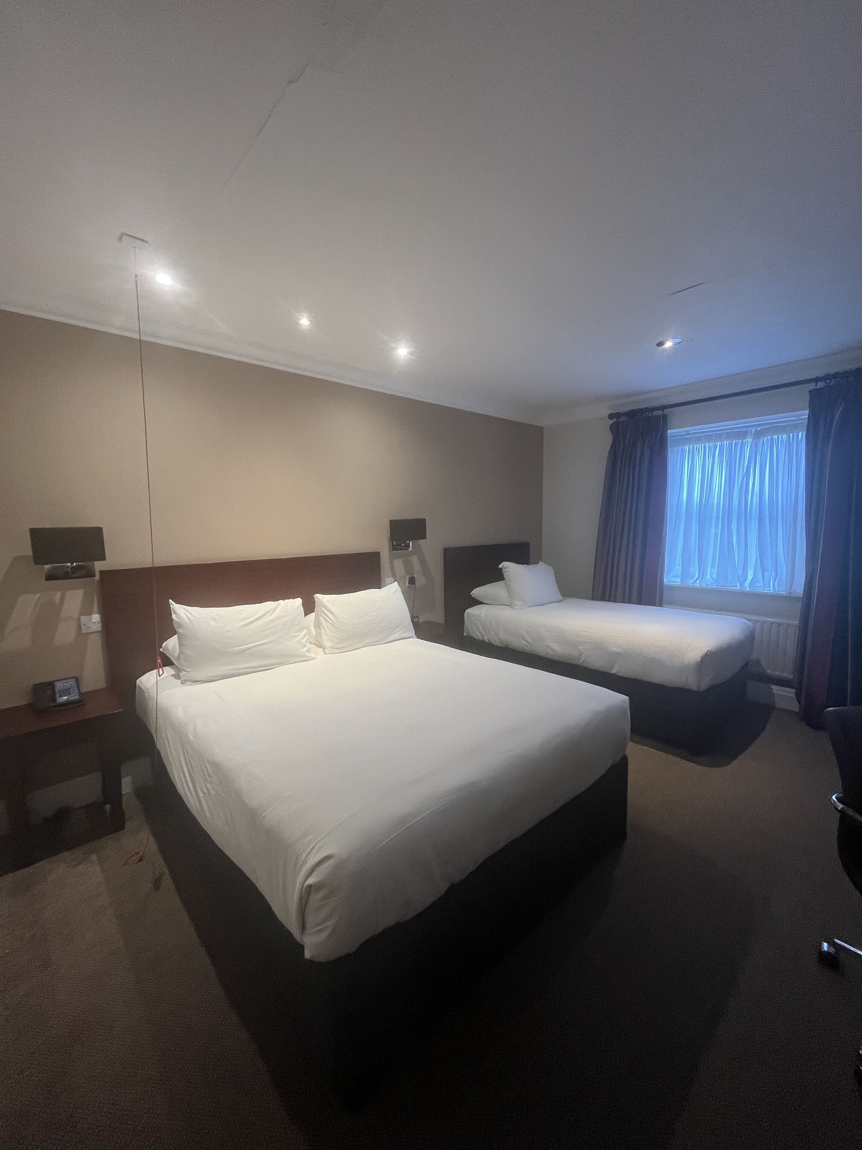 Ein Hotelzimmer mit zwei Betten, einem Schreibtisch, einem Stuhl und einem fensterbepflanzten Fenster.