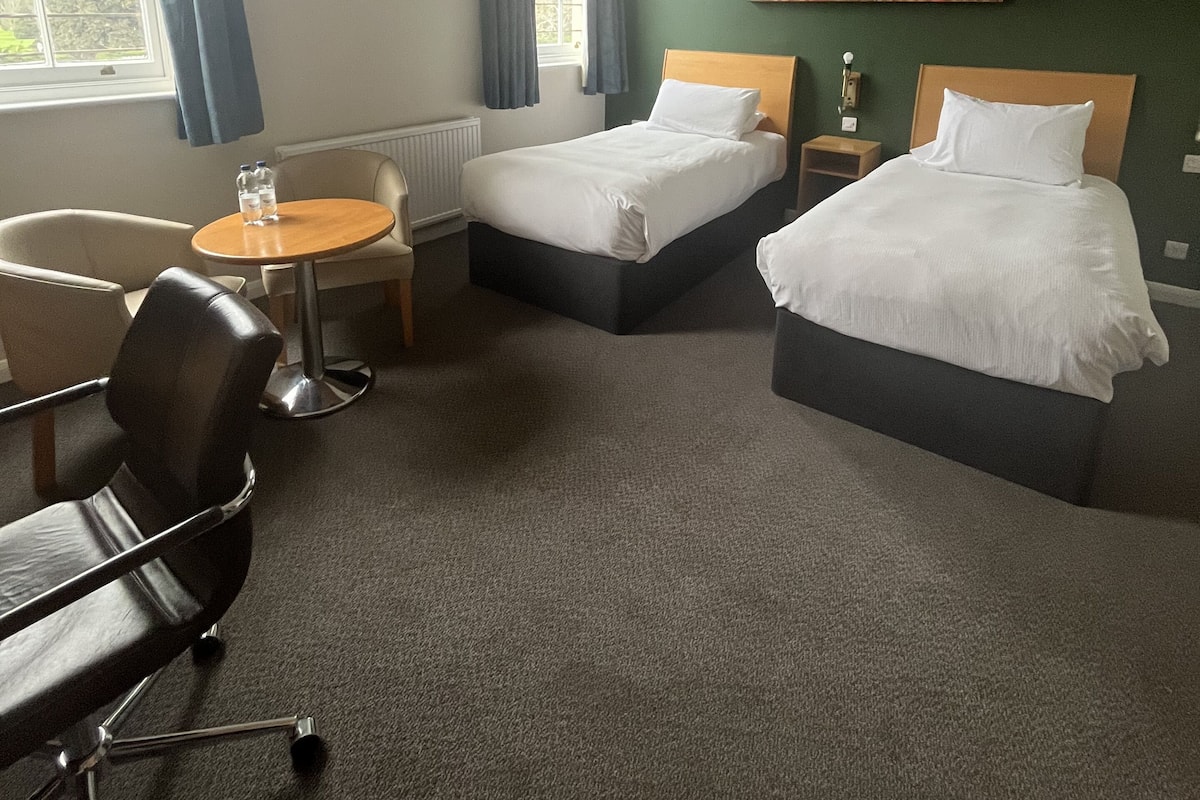 A hotel room with two beds, a round table, a chair, and a window with curtains.
