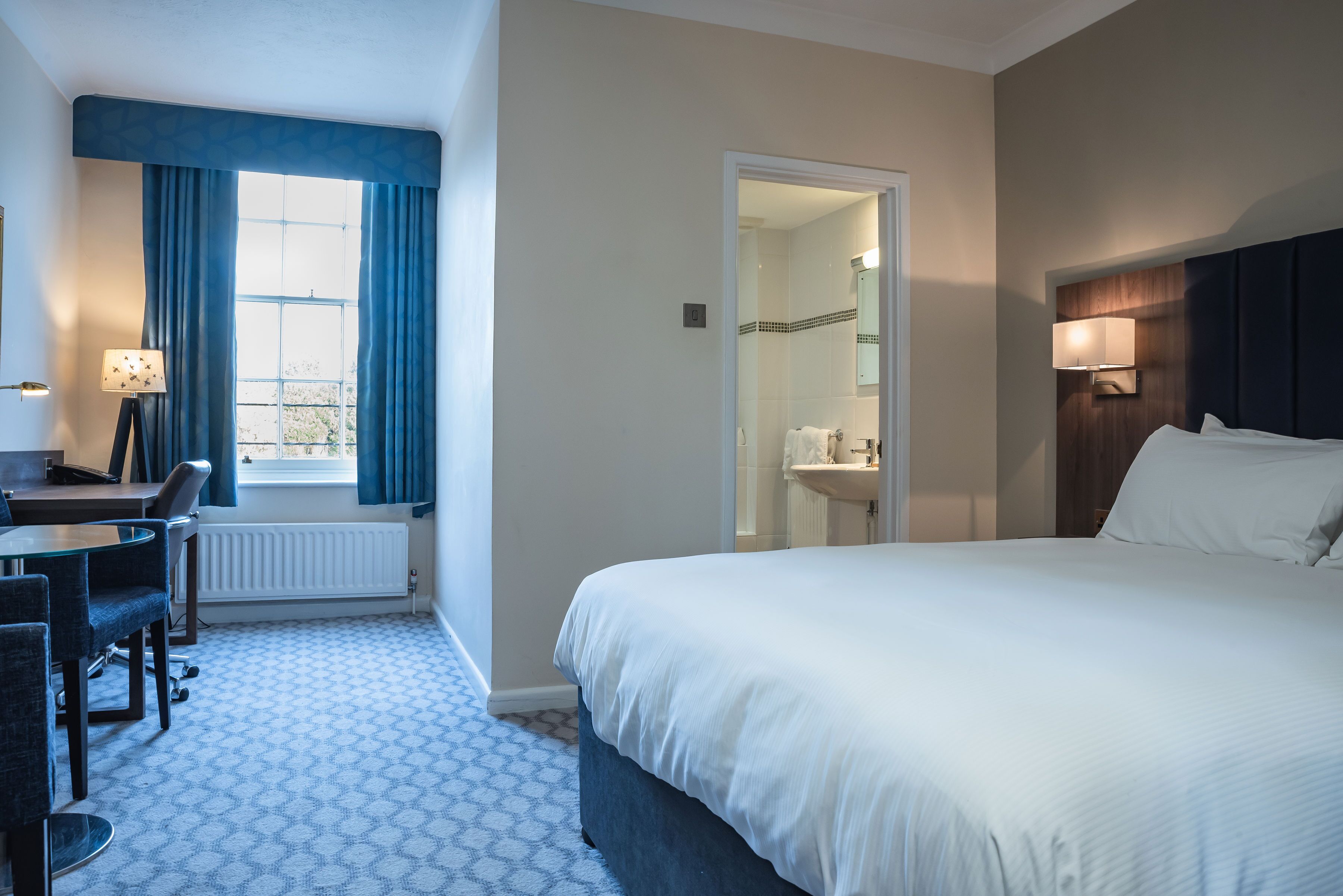 Une chambre d’hôtel avec un grand lit, un bureau avec une chaise, une fenêtre avec des rideaux, une salle de bain avec un miroir et un lavabo, et une lampe fixée au mur.