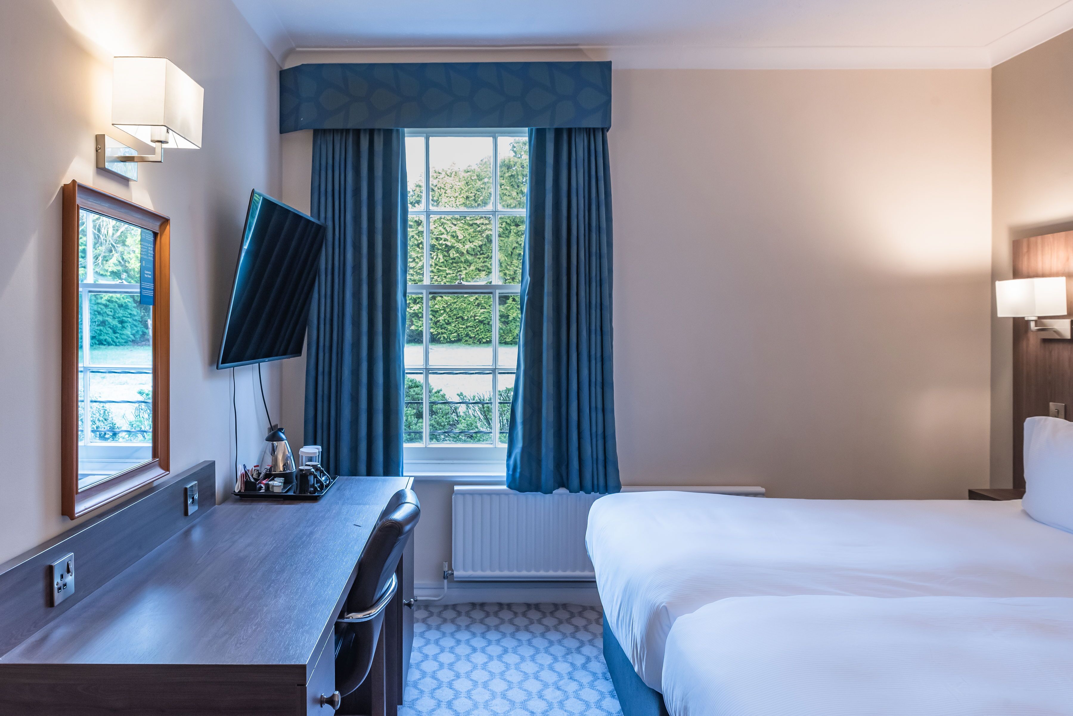 A hotel room with a bed, a desk with a chair, a TV, and a window with curtains.