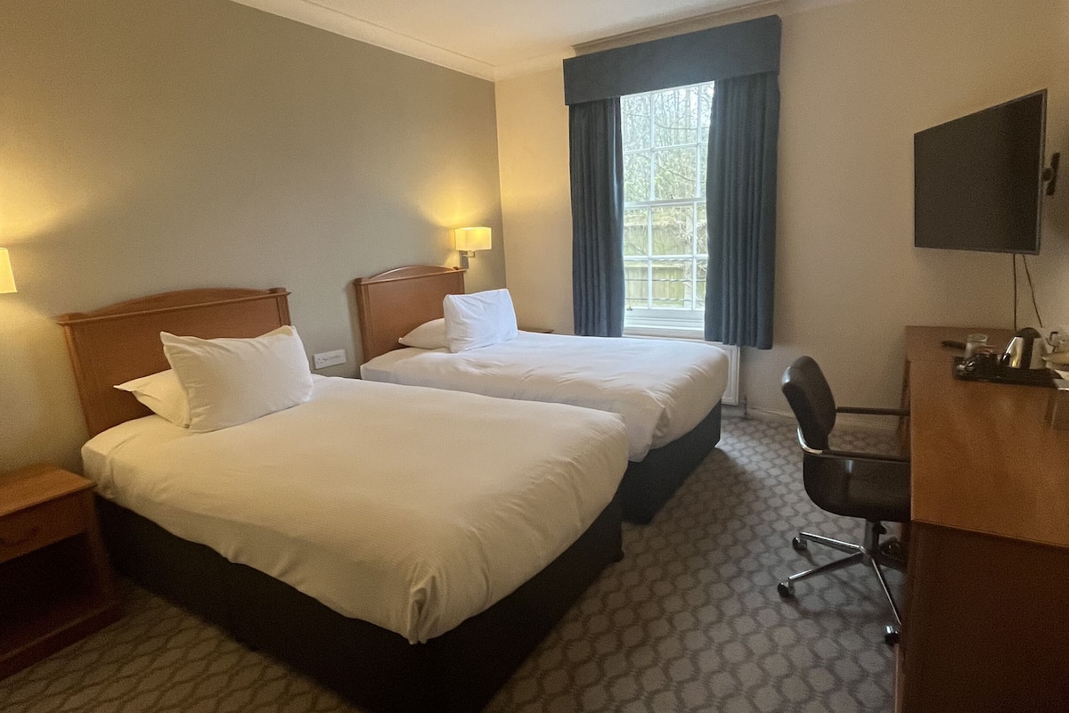 A hotel room with two beds, a desk, a chair, a TV, and a window with curtains.
