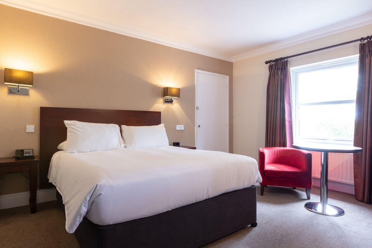 A hotel room with a large bed, two bedside lamps, a red chair, and a window with curtains.