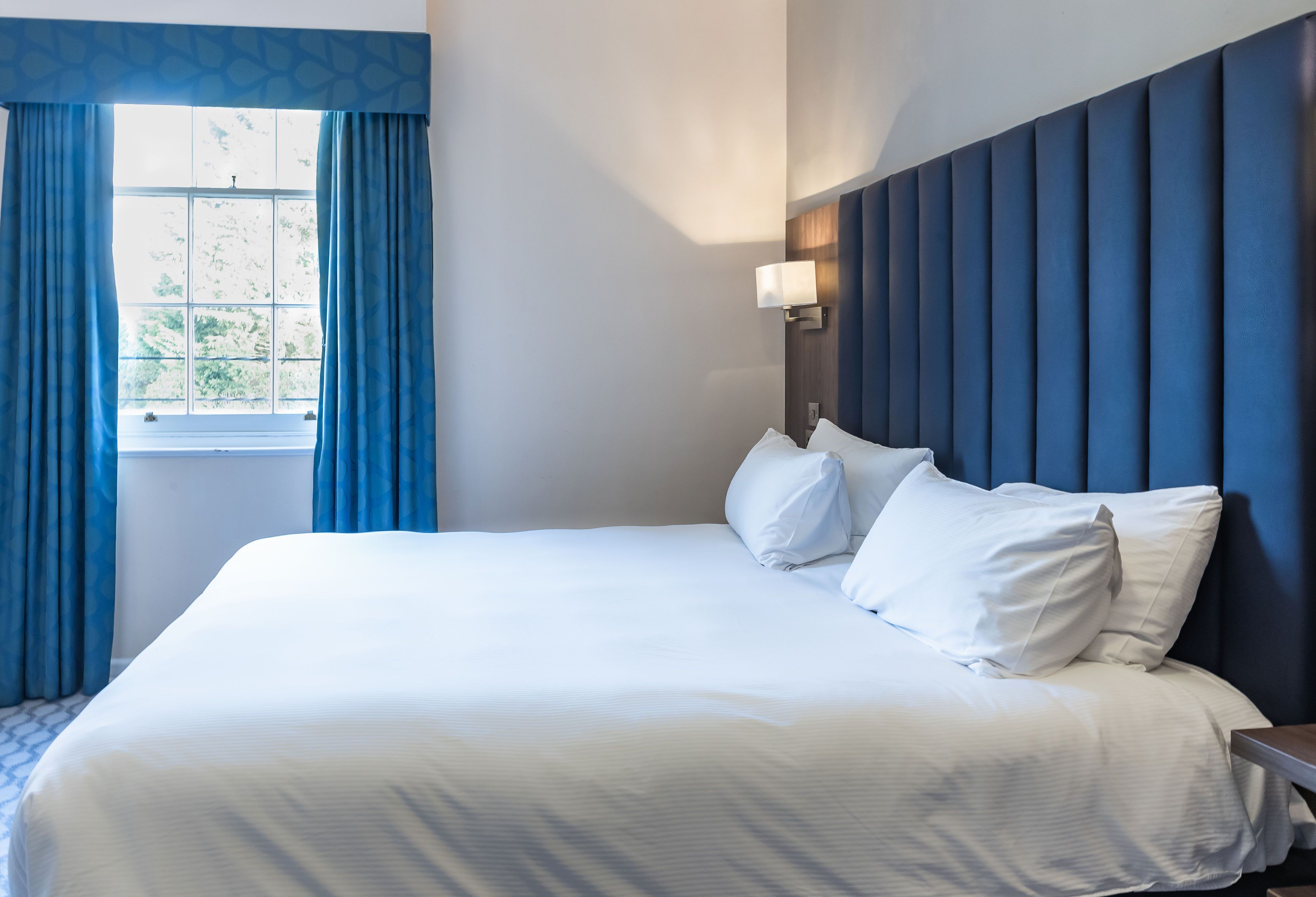 A hotel room with a large bed, a window with blue curtains, a wall-mounted lamp, and a blue headboard.