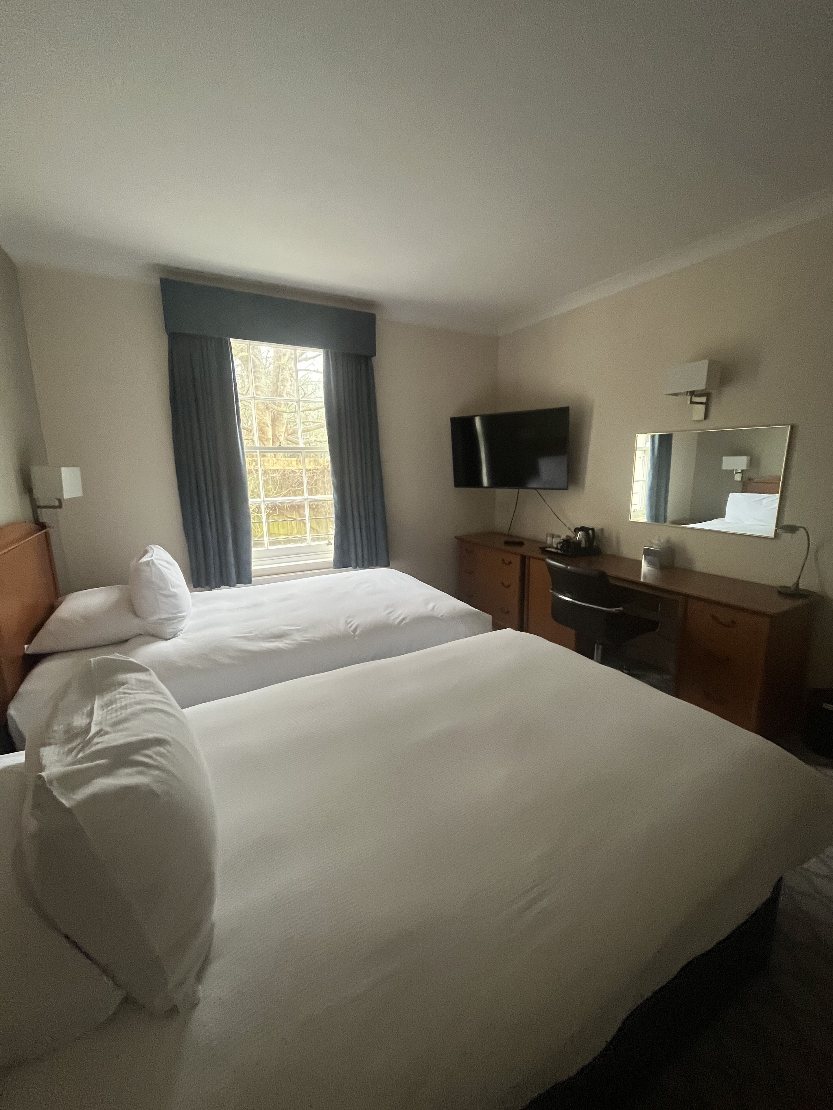 A hotel room with two beds, a desk, a chair, a television, and a window with curtains.