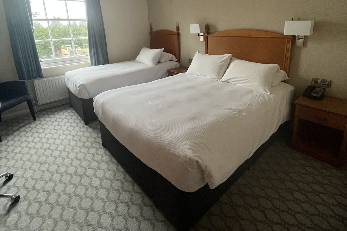 A hotel room with two beds, a window with curtains, a bedside table with a lamp, and a radiator.