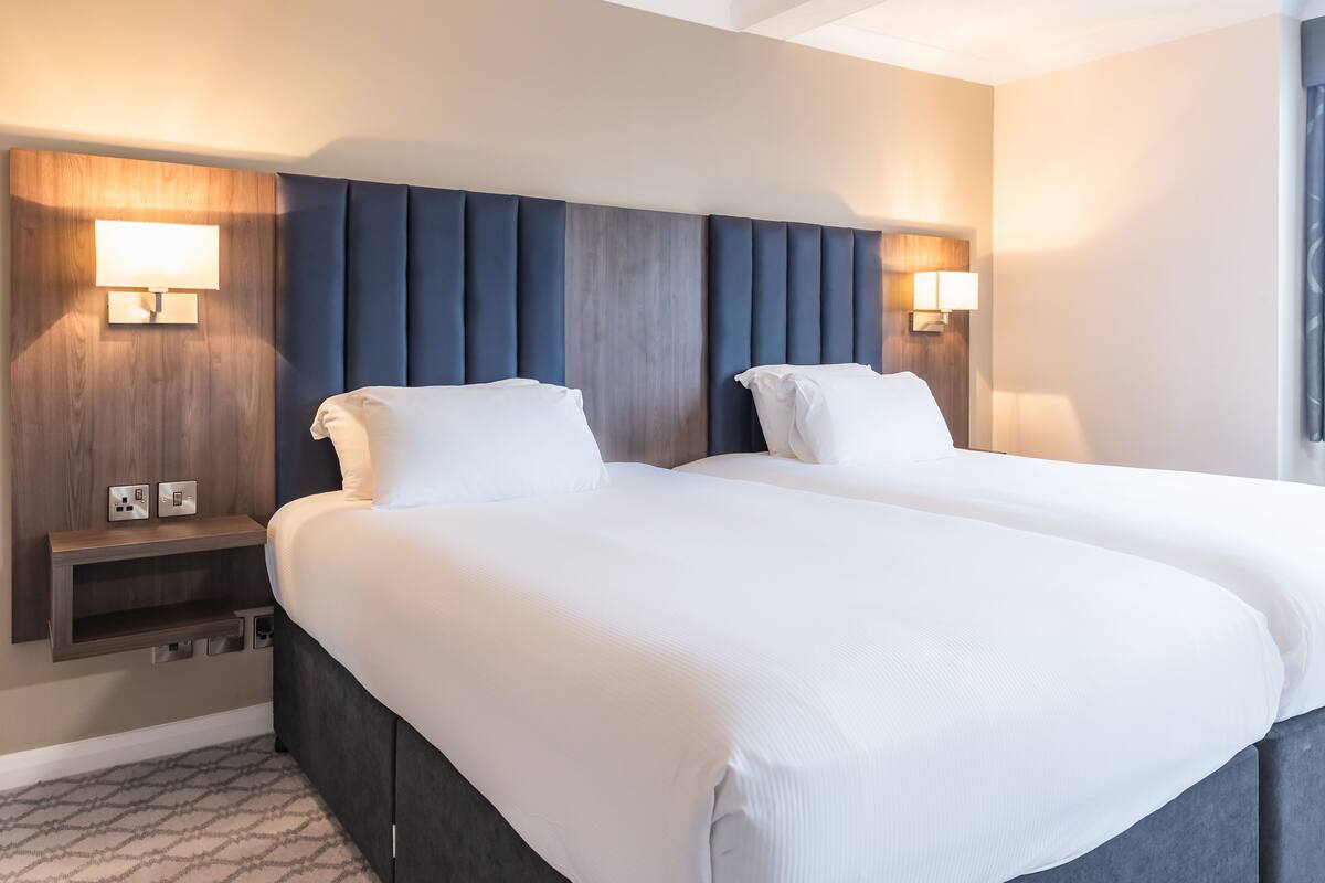 A hotel room with two beds, a wooden headboard, bedside lamps, and a patterned carpet.