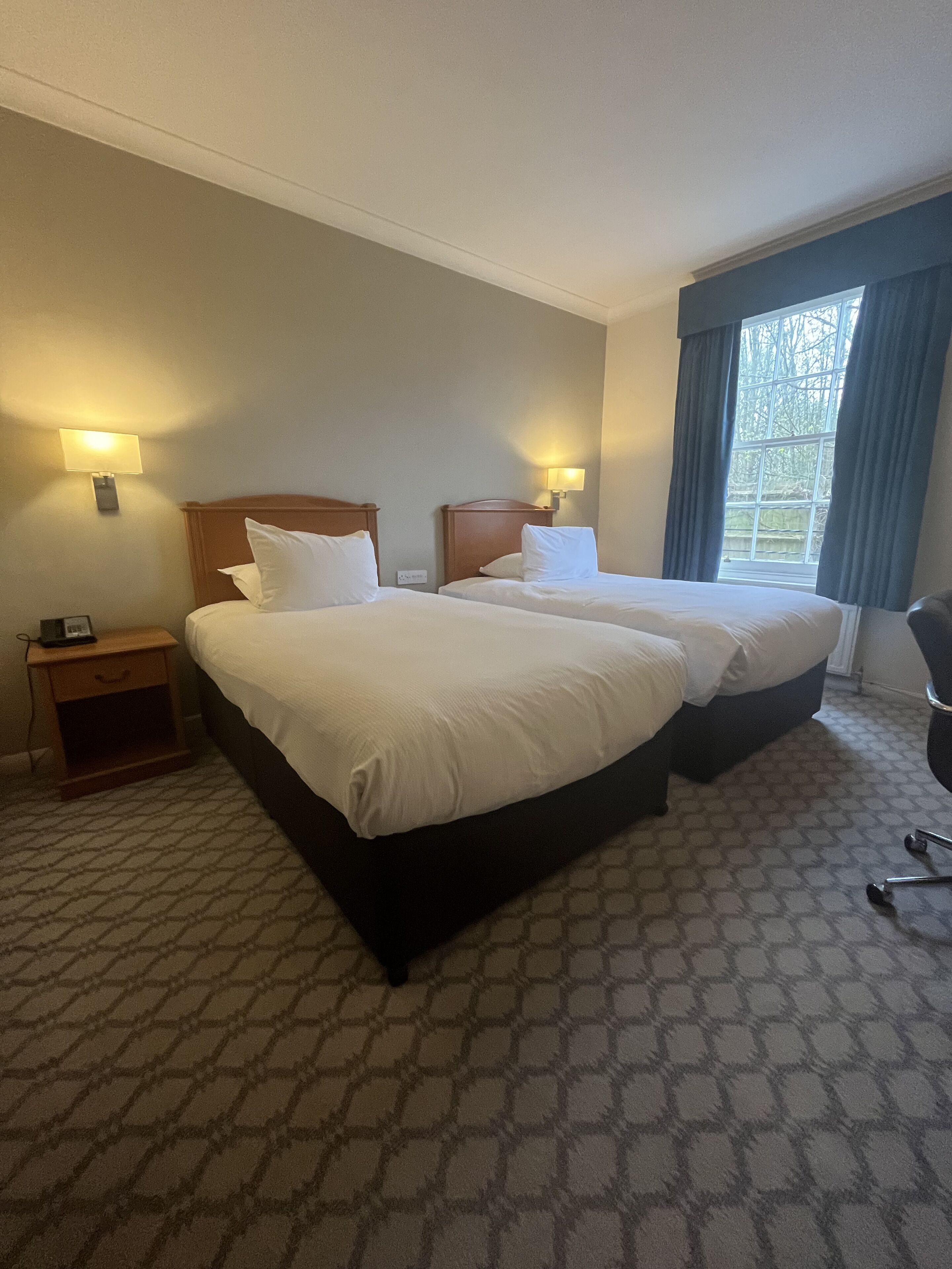 A hotel room with two beds, a nightstand, a chair, and a window with curtains.