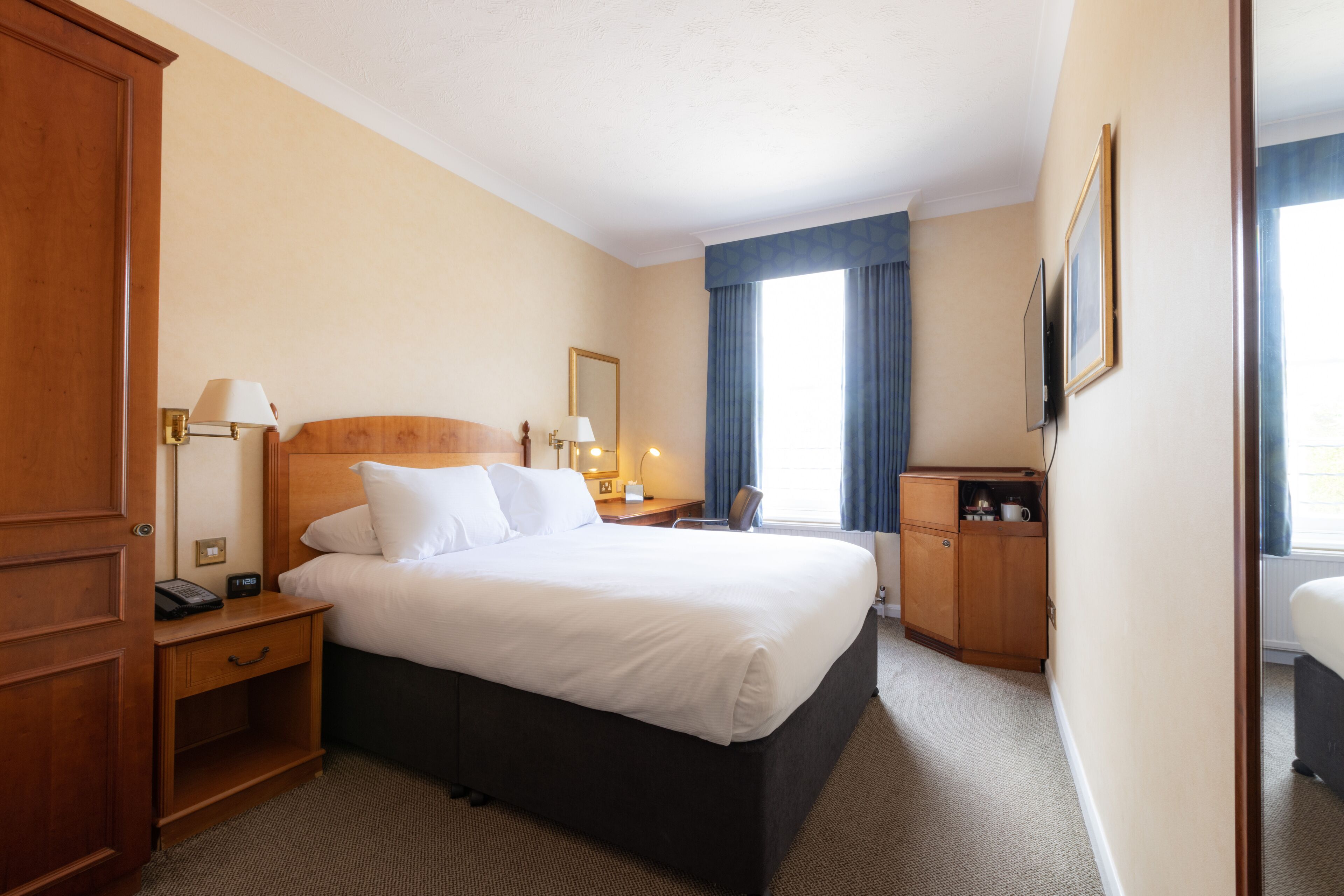 A hotel room with a large bed, bedside tables, a desk, a chair, and a television.