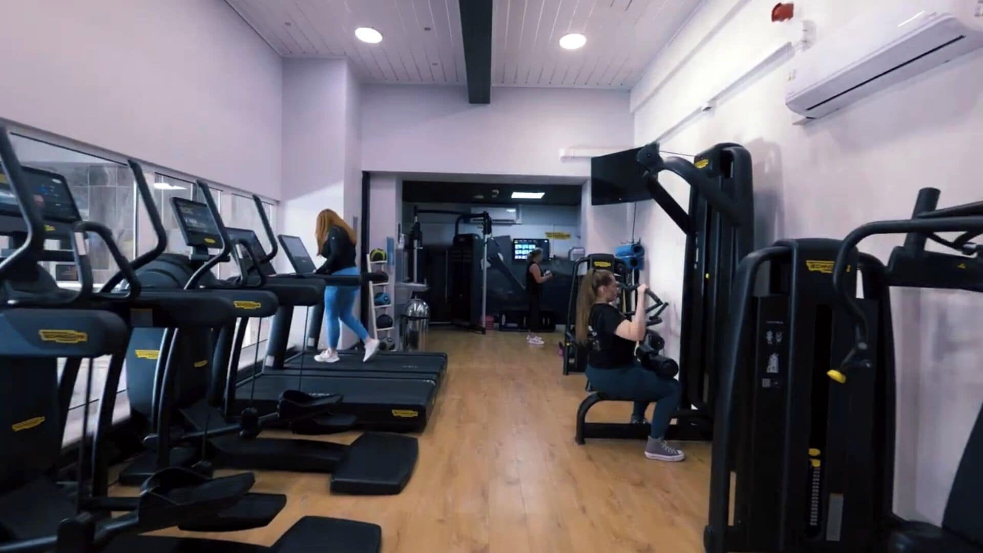 Un gimnasio con cintas de correr, máquinas de pesas y suelo de madera.