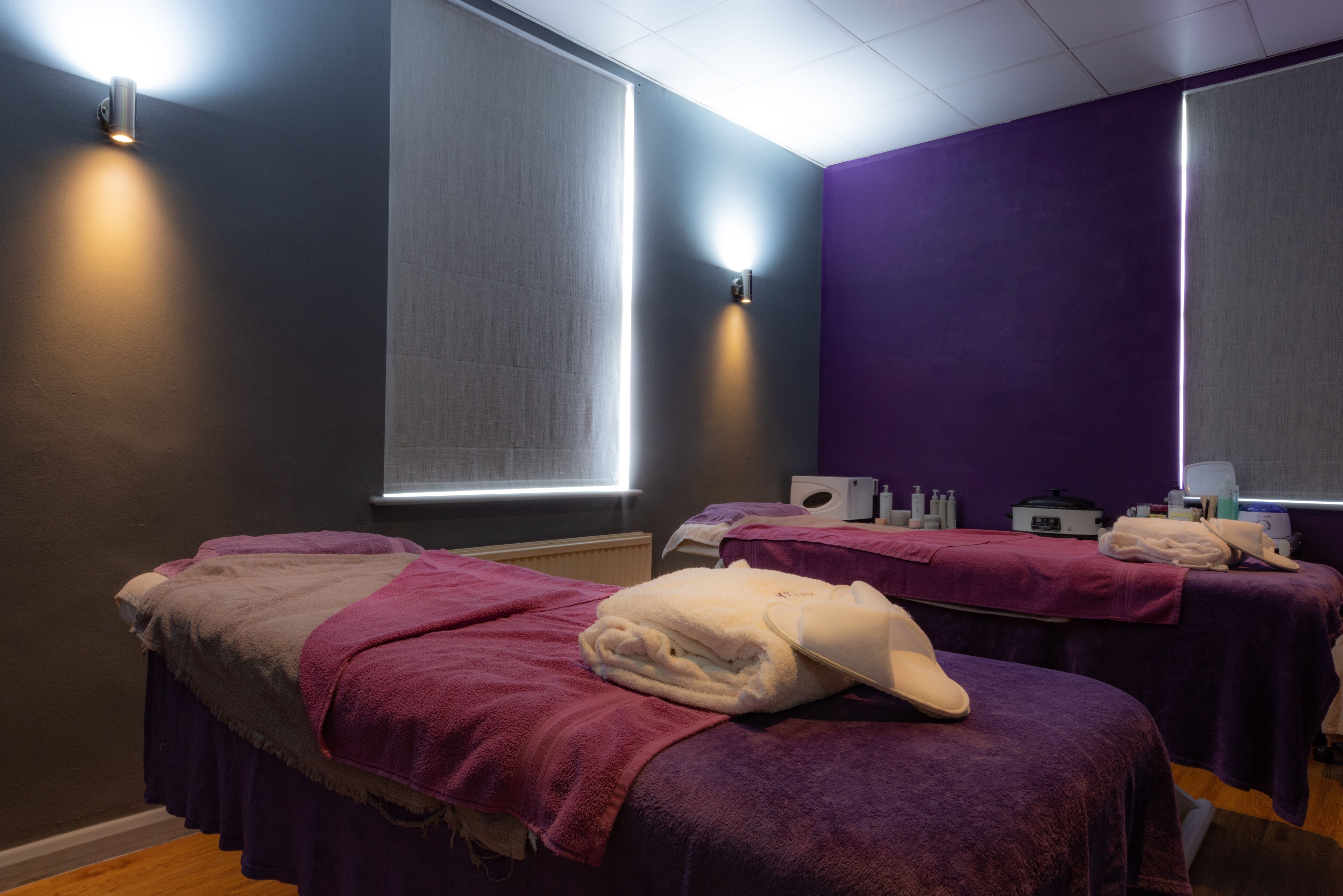 A room with two massage tables, each with a purple towel and a white cap. There are wall-mounted lights and a window with blinds.