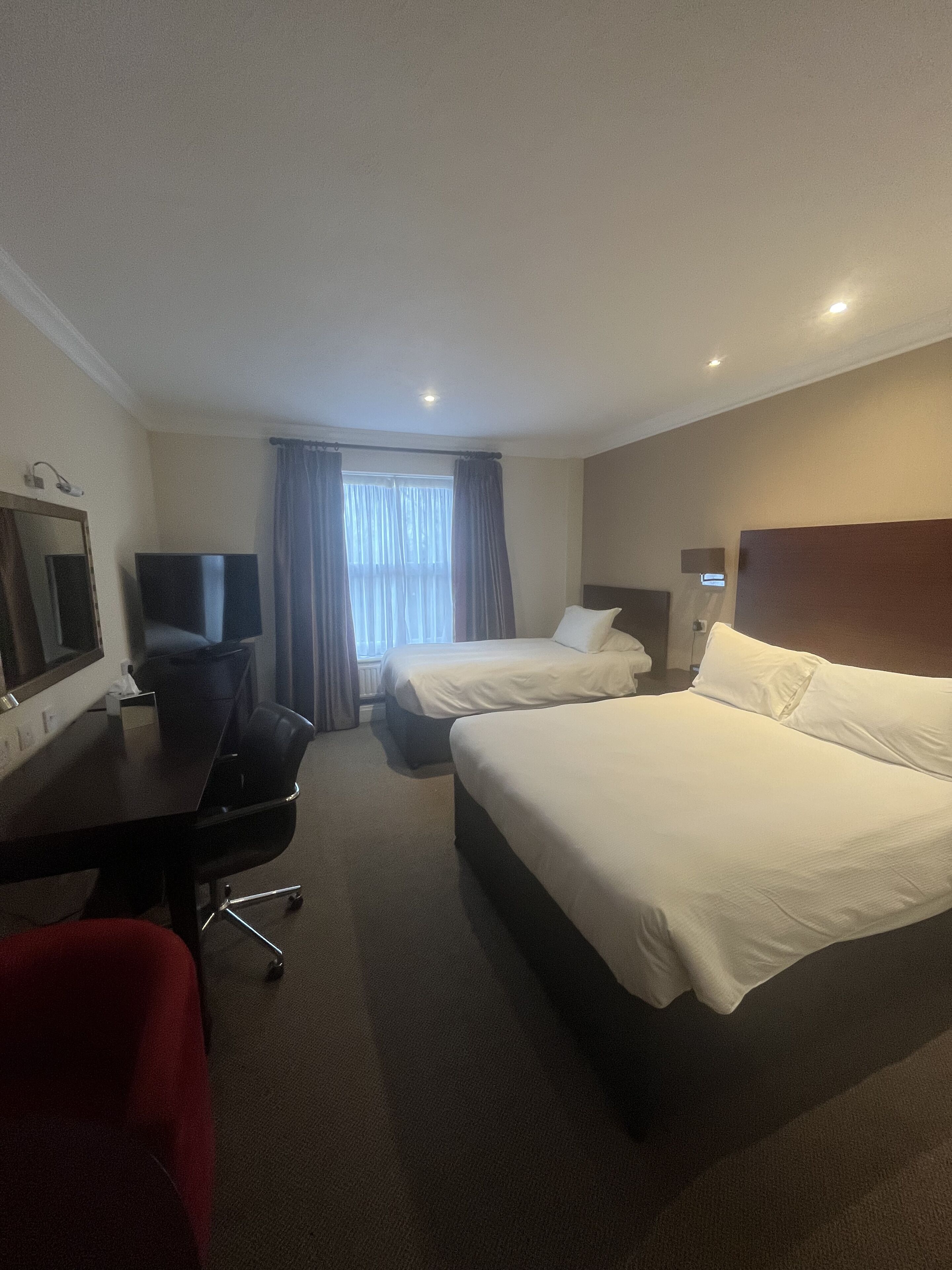 Une chambre d’hôtel avec deux lits, un bureau, une chaise, une télévision et une fenêtre avec des rideaux.