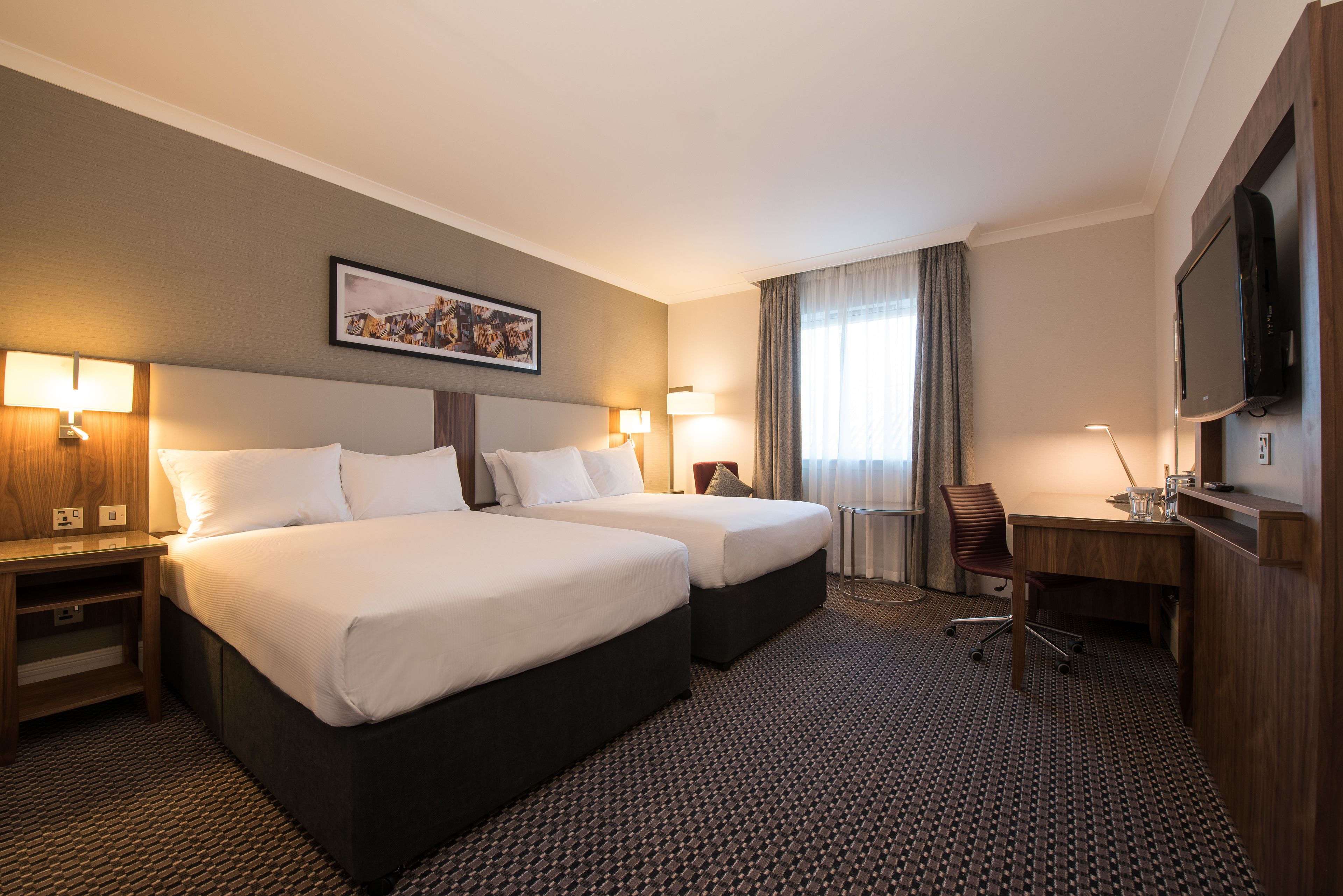 A hotel room with two beds, a desk, a chair, a TV, and a picture on the wall.