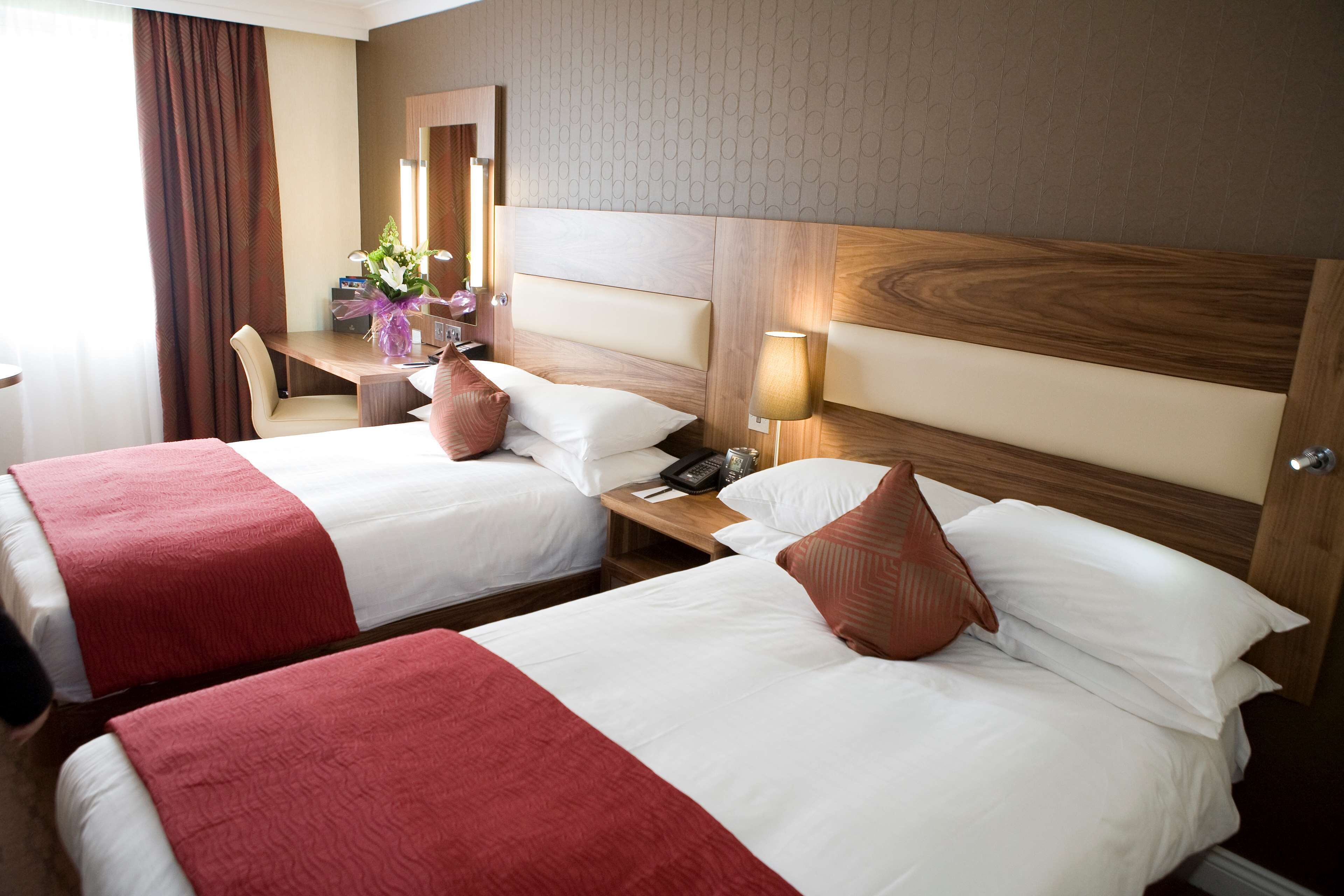 Habitación de hotel con dos camas, cada una con cubrecama rojo y blanco, cabecero de madera, mesita de noche con lámpara y espejo en la pared.
