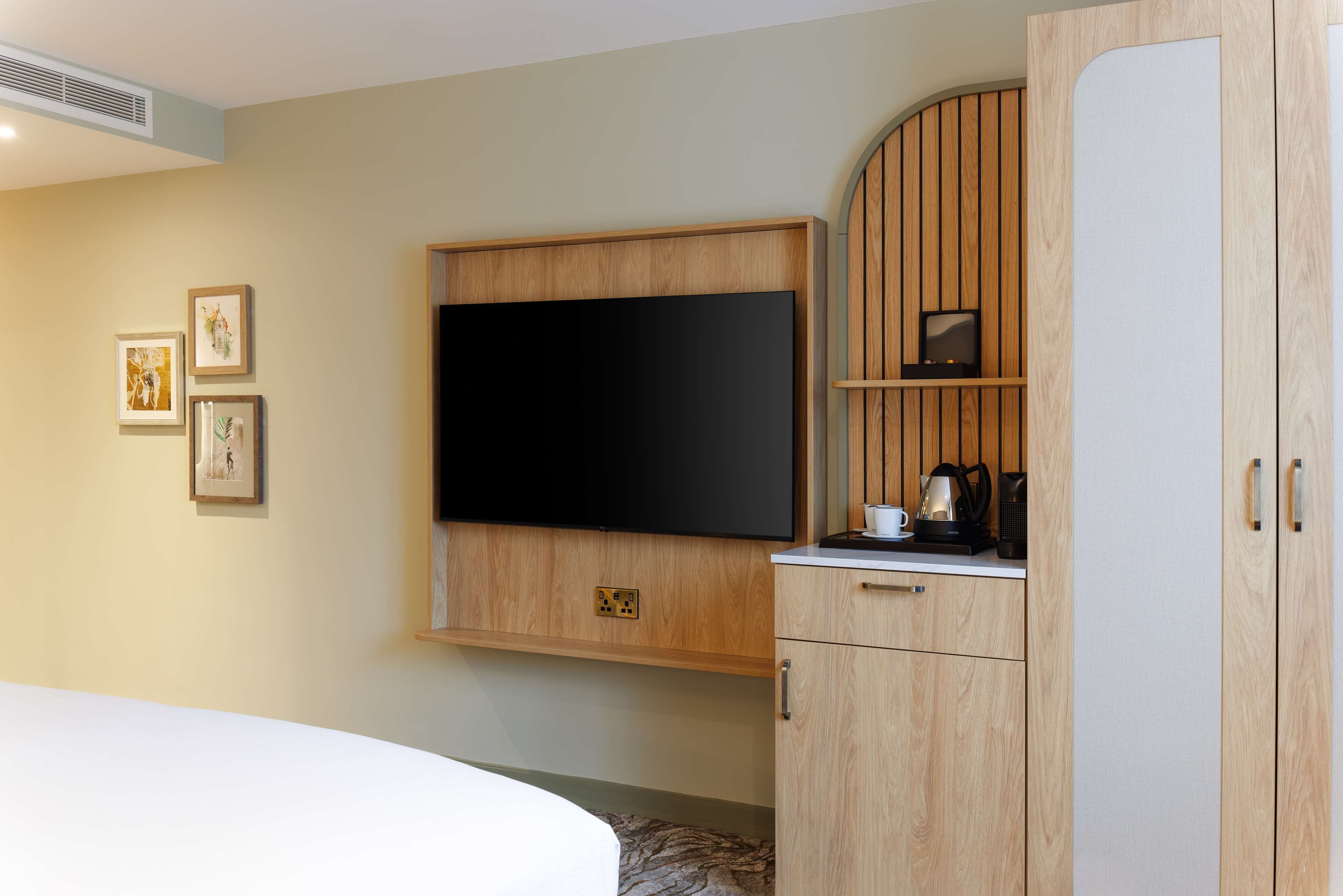 Une chambre d’hôtel équipée d’un lit, d’un téléviseur à écran plat fixé au mur, d’armoires en bois et d’une petite étagère avec une bouilloire et des tasses.