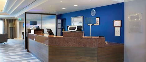 Un espace d’accueil avec un mur bleu, un comptoir de réception, un écran numérique et un panneau d’information sur la sécurité incendie.