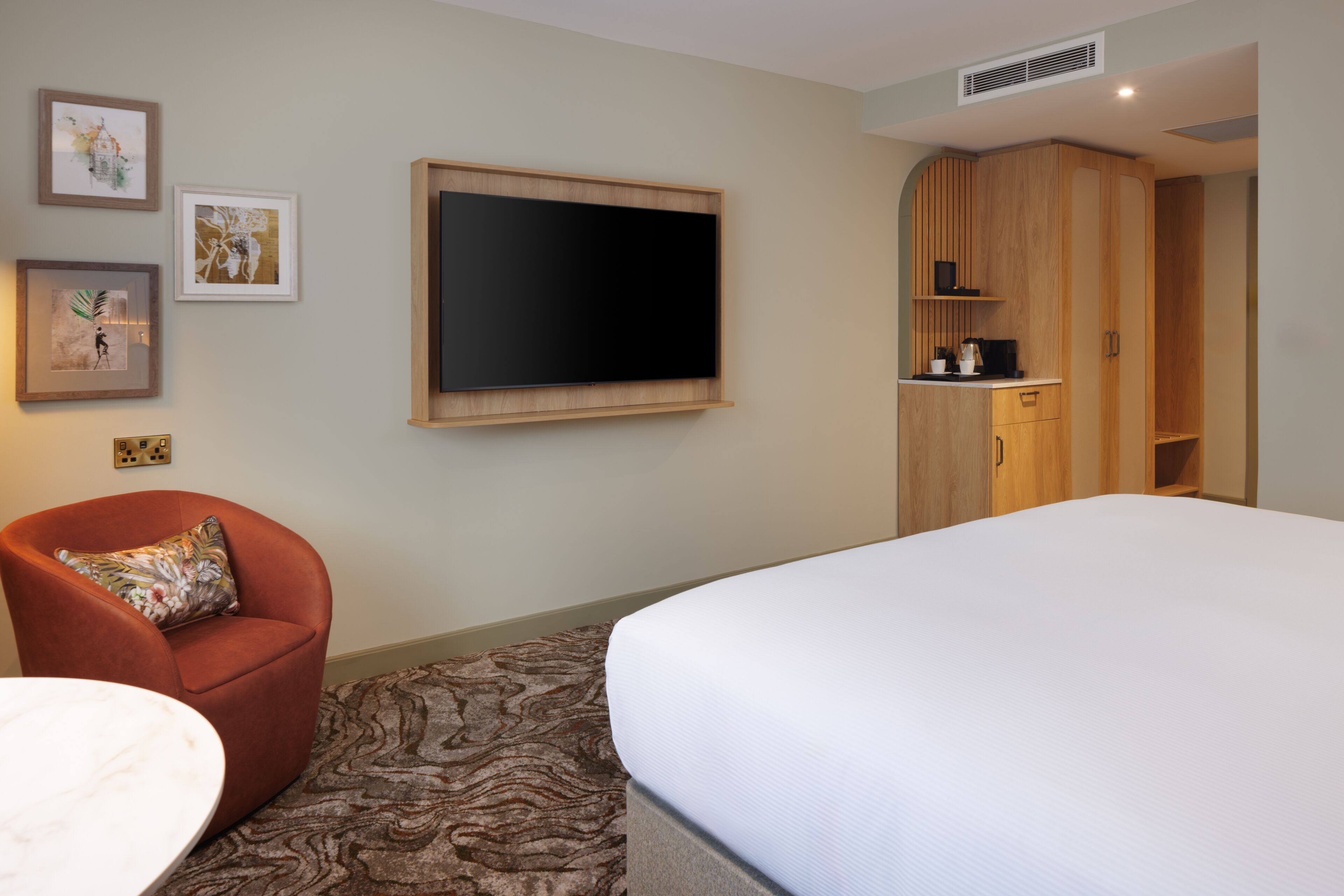 A hotel room with a bed, a TV mounted on the wall, a wooden wardrobe, a red armchair, and a patterned carpet.