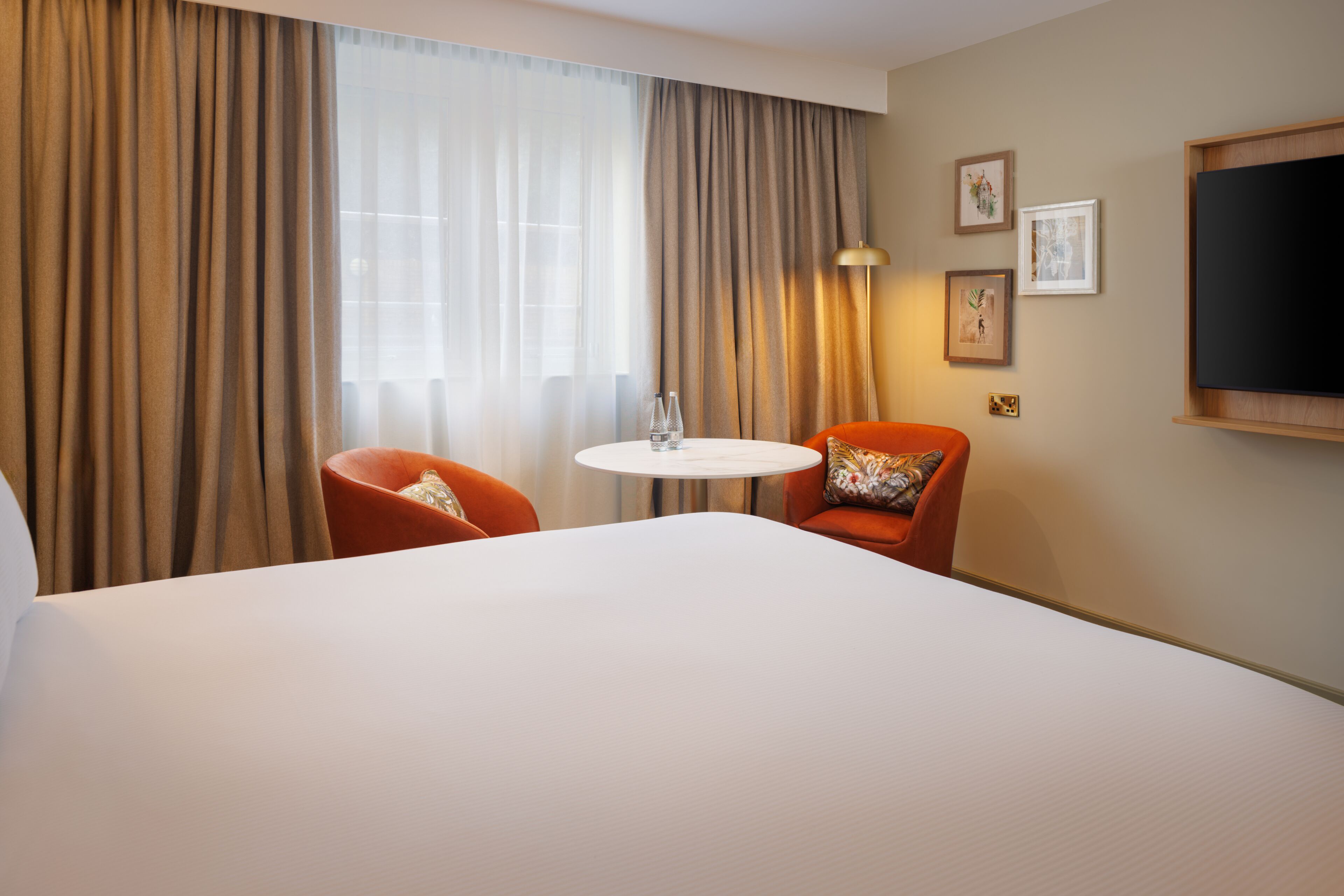 A hotel room with a large bed, two orange chairs, a small round table with a glass, and a TV mounted on the wall.
