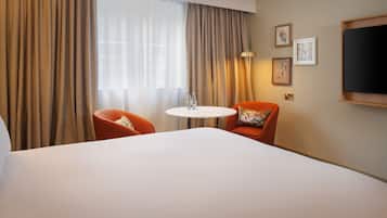 A hotel room with a large bed, two orange chairs, a small round table with a glass, and a TV mounted on the wall.