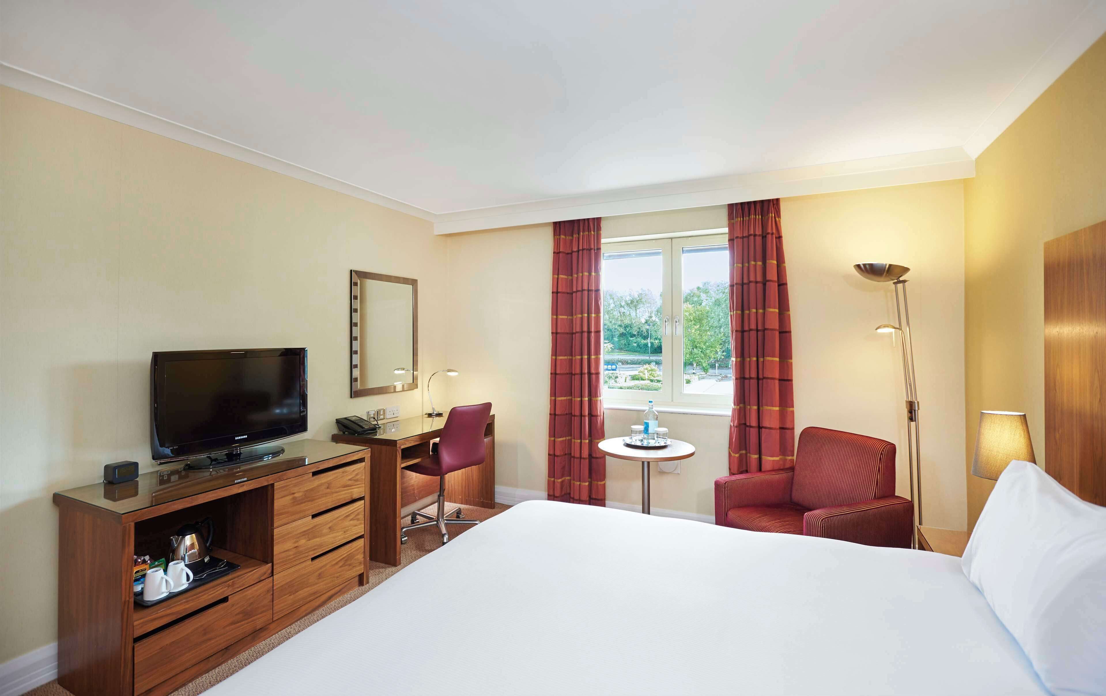 A hotel room with a bed, a desk with a television, a chair, a small table, a lamp, and a window with curtains.