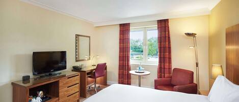 Habitación de hotel con cama, escritorio con televisor, silla, mesita, lámpara y ventana con cortinas.