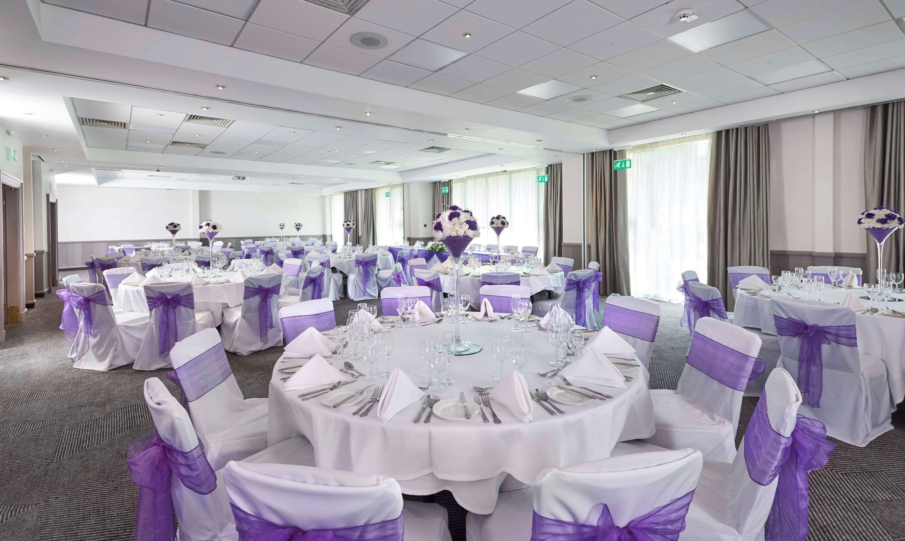 Une salle de banquet avec des tables rondes dressées pour un événement formel, avec des nappes blanches et des rubans violets sur les chaises.
