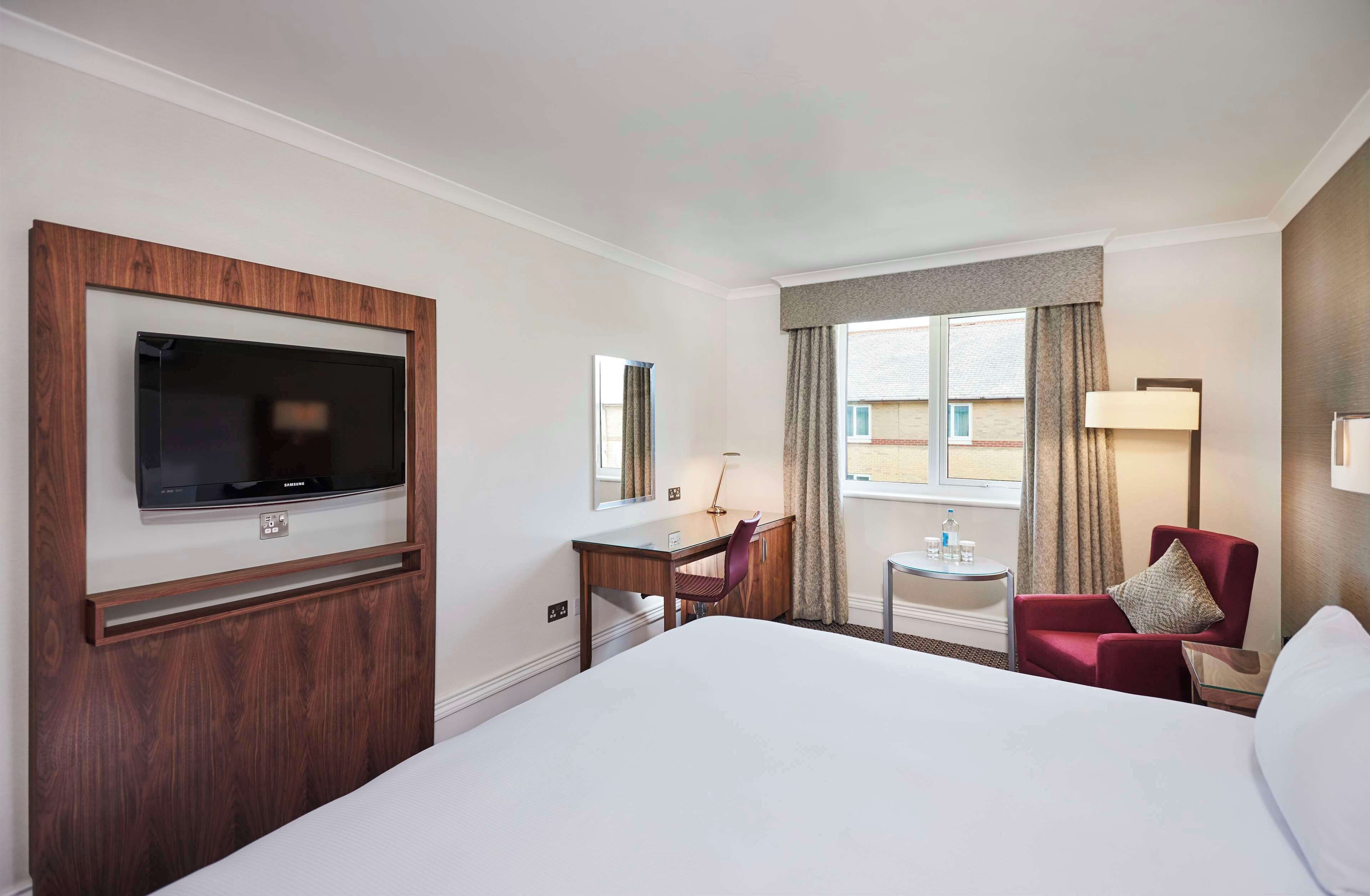 A hotel room with a large bed, a desk with a chair, a television mounted on a wooden panel, a small table with a lamp, and a window with curtains.