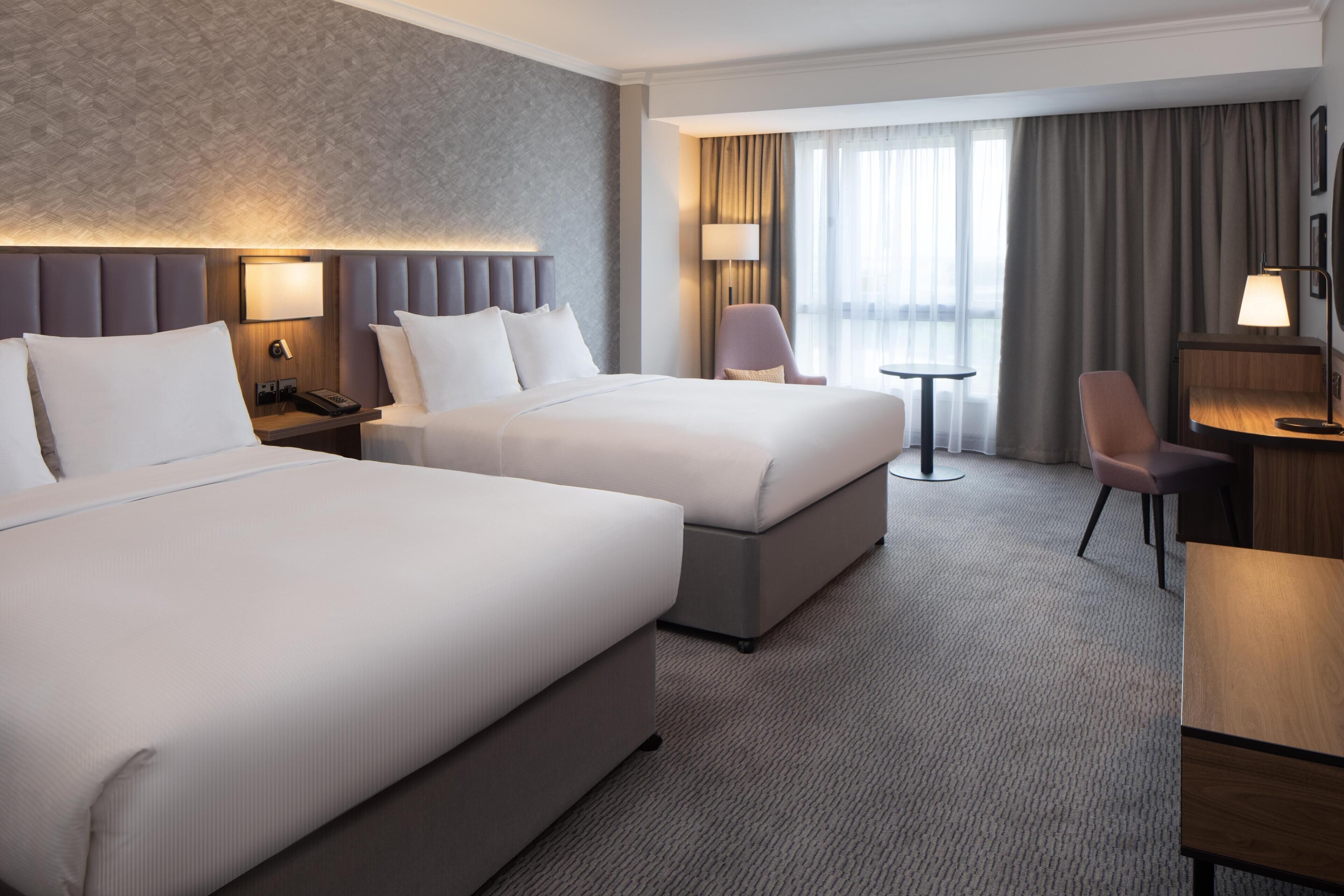 Hotellihuone, jossa on kaksi sänkyä, työpöytä, tuoli, lamppu ja näkymä ulos.