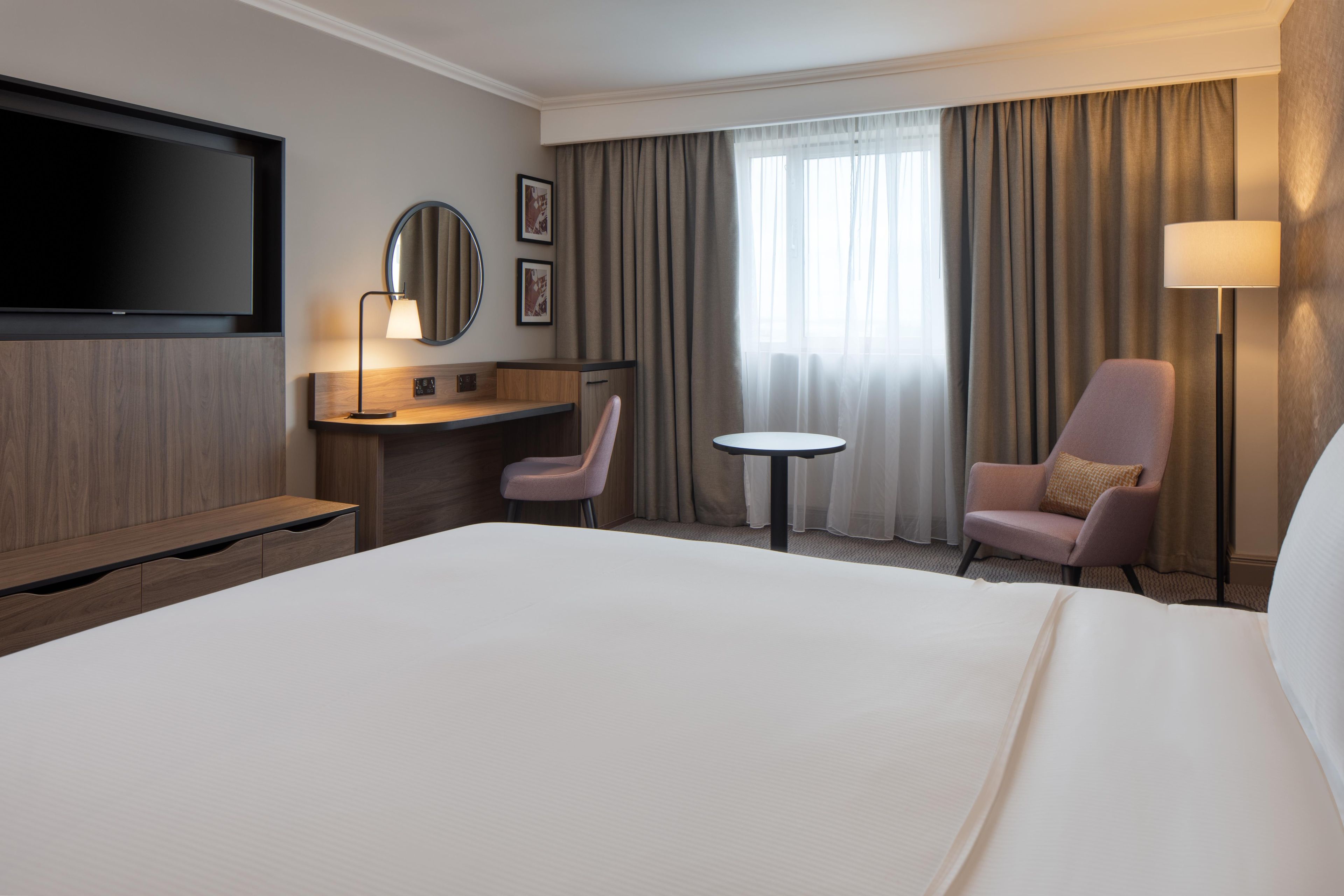 Een hotelkamer met een groot bed, een bureau met stoel, een kleine tafel, een aan de muur bevestigde televisie en een raam met gordijnen.