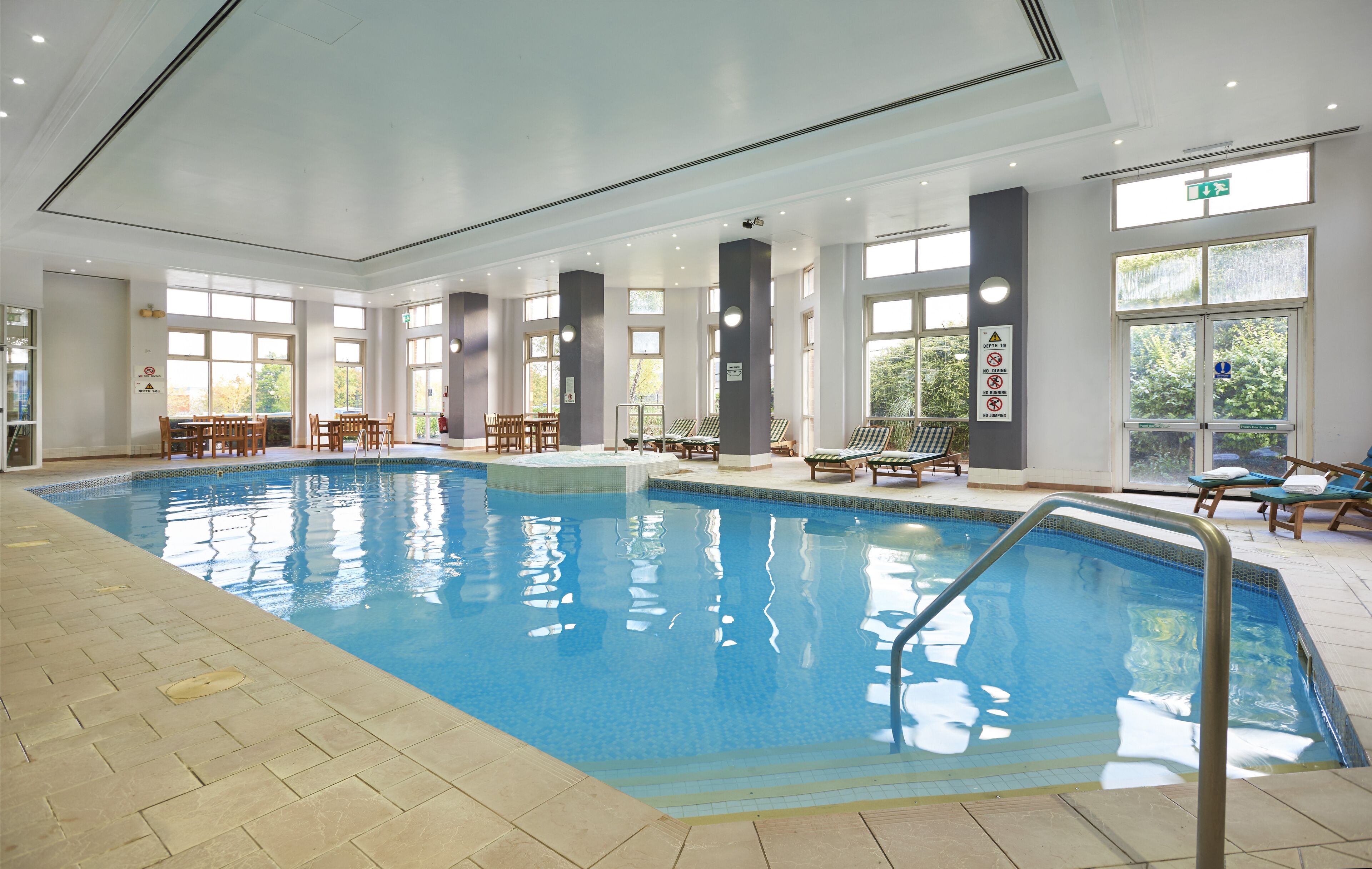 An indoor swimming pool with clear water, surrounded by lounge chairs and tables, large windows, and a tiled floor.
