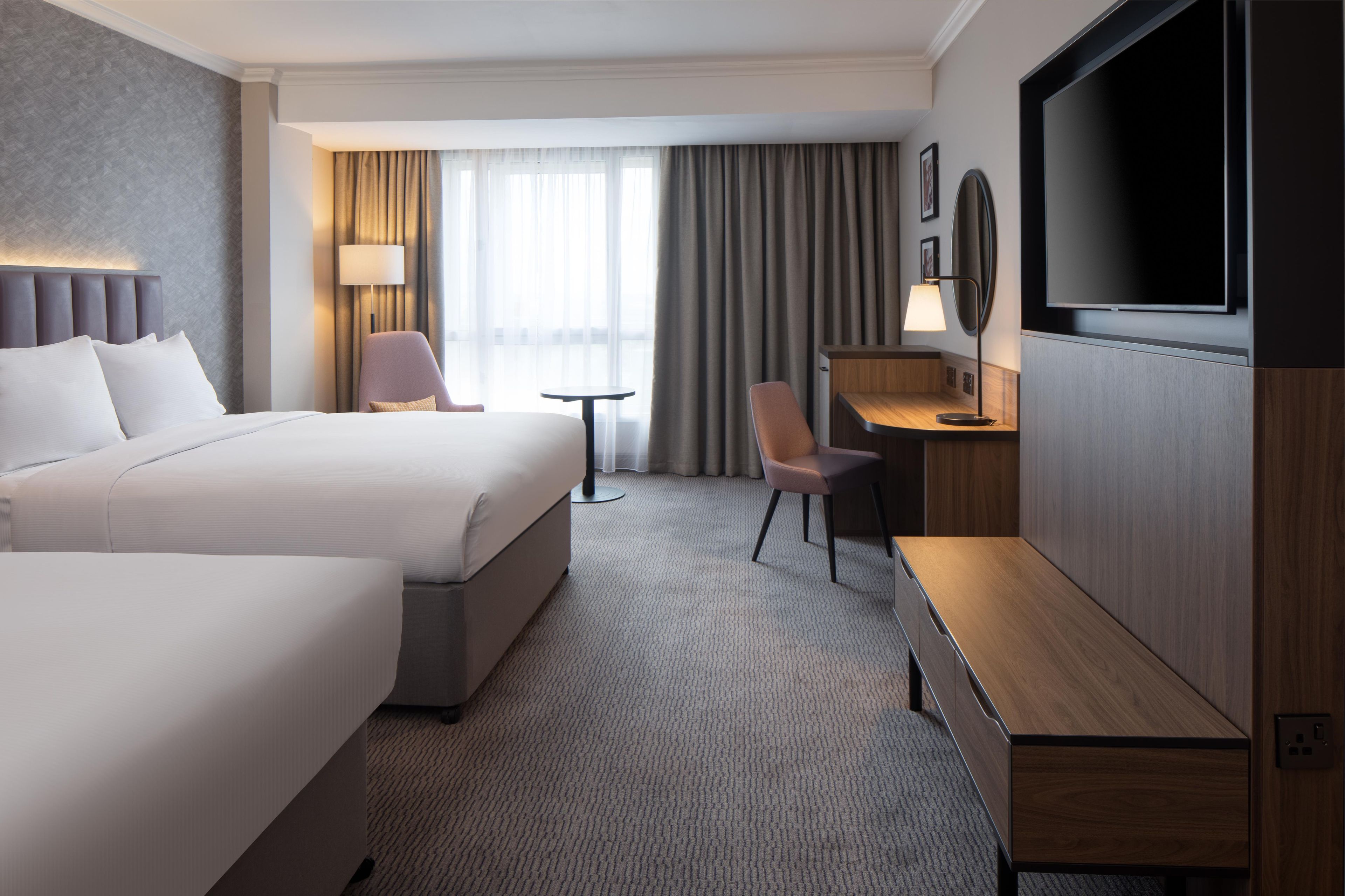 A hotel room with a large bed, a desk, a chair, a TV, and a window with curtains.