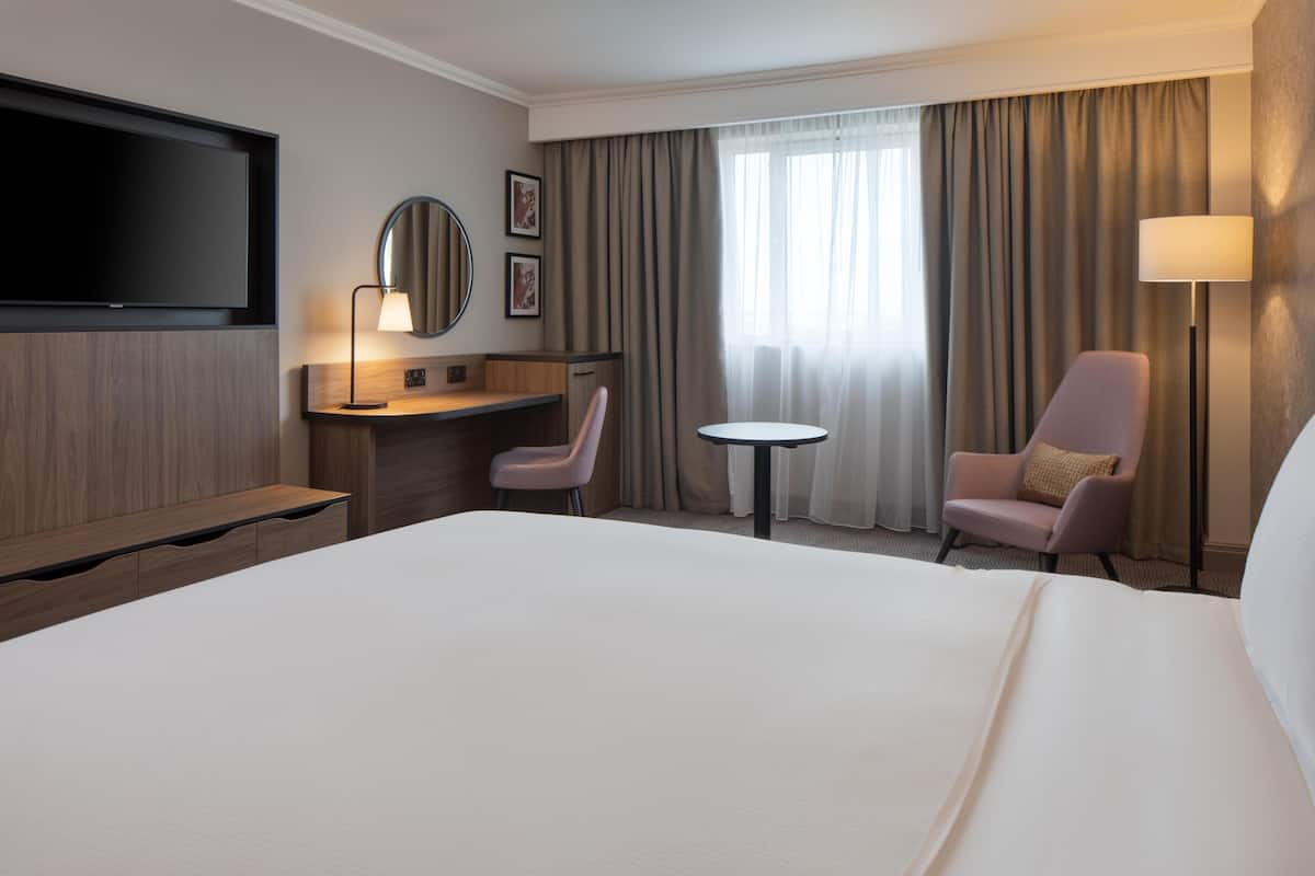 Hotelkamer met een groot bed, bureau met stoel, een klein tafeltje, een aan de muur bevestigde televisie en een raam met gordijnen.
