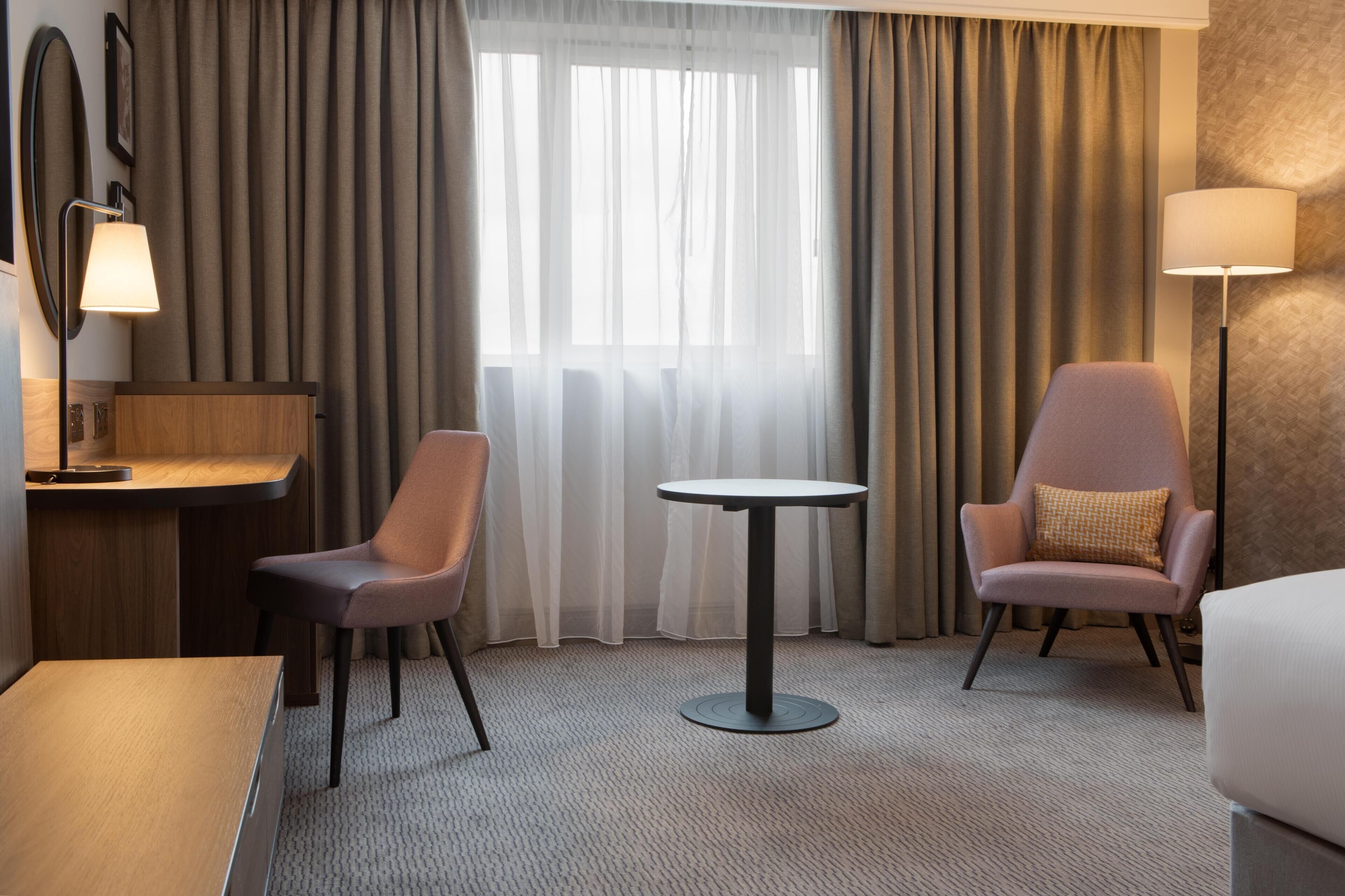 Une chambre d’hôtel équipée d’un bureau, de deux chaises, d’une table ronde, d’une lampe et d’une grande fenêtre avec des rideaux.