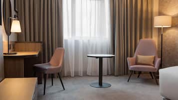 Une chambre d’hôtel équipée d’un bureau, de deux chaises, d’une table ronde, d’une lampe et d’une grande fenêtre avec des rideaux.