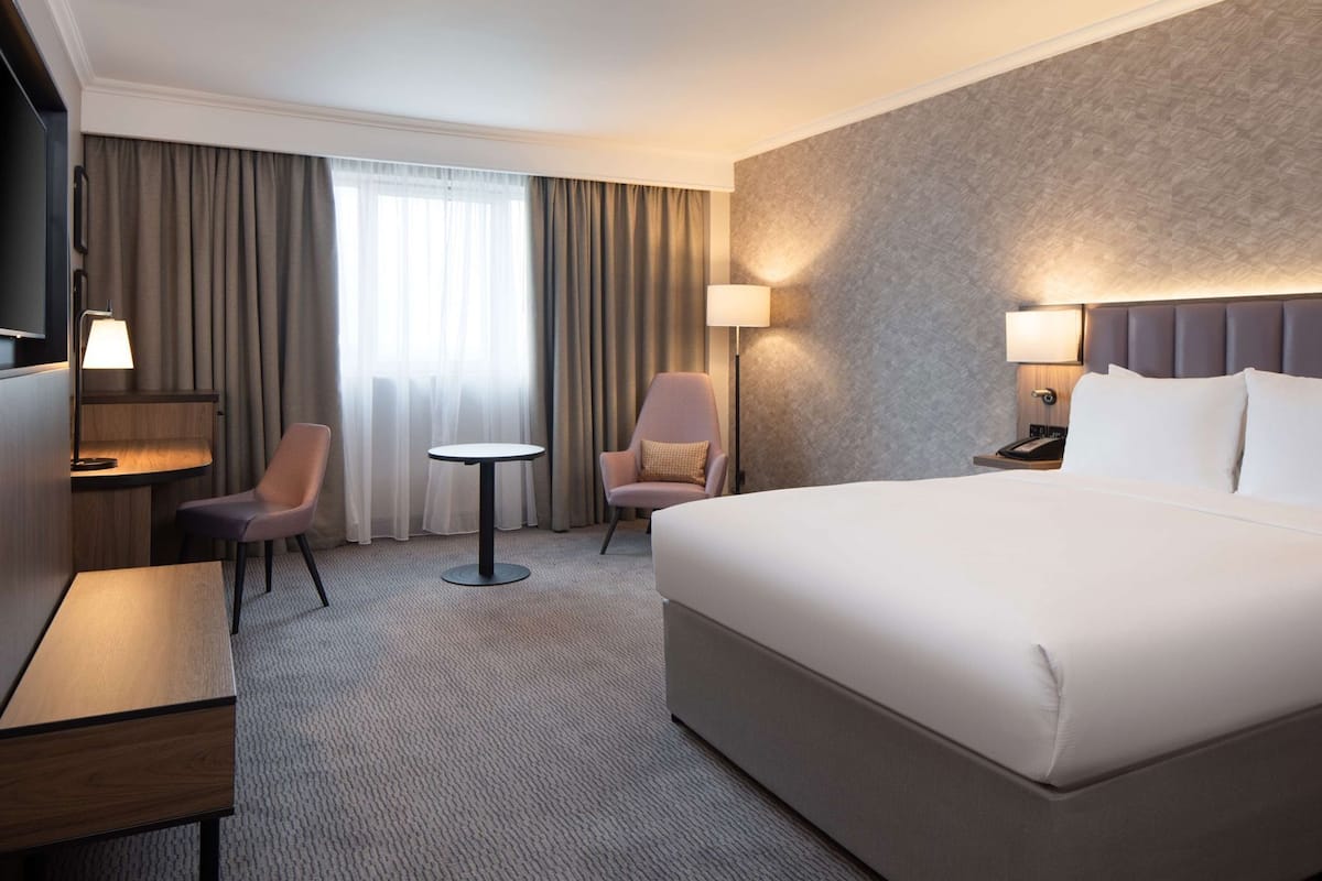 Ein Hotelzimmer mit einem großen Bett, zwei Sesseln, einem kleinen Tisch, einem Schreibtisch und einem Fernseher.