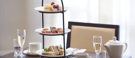 Un soporte de tres niveles para postres con diversas pastas y sándwiches, una tetera, tazas y vasos sobre una mesa.