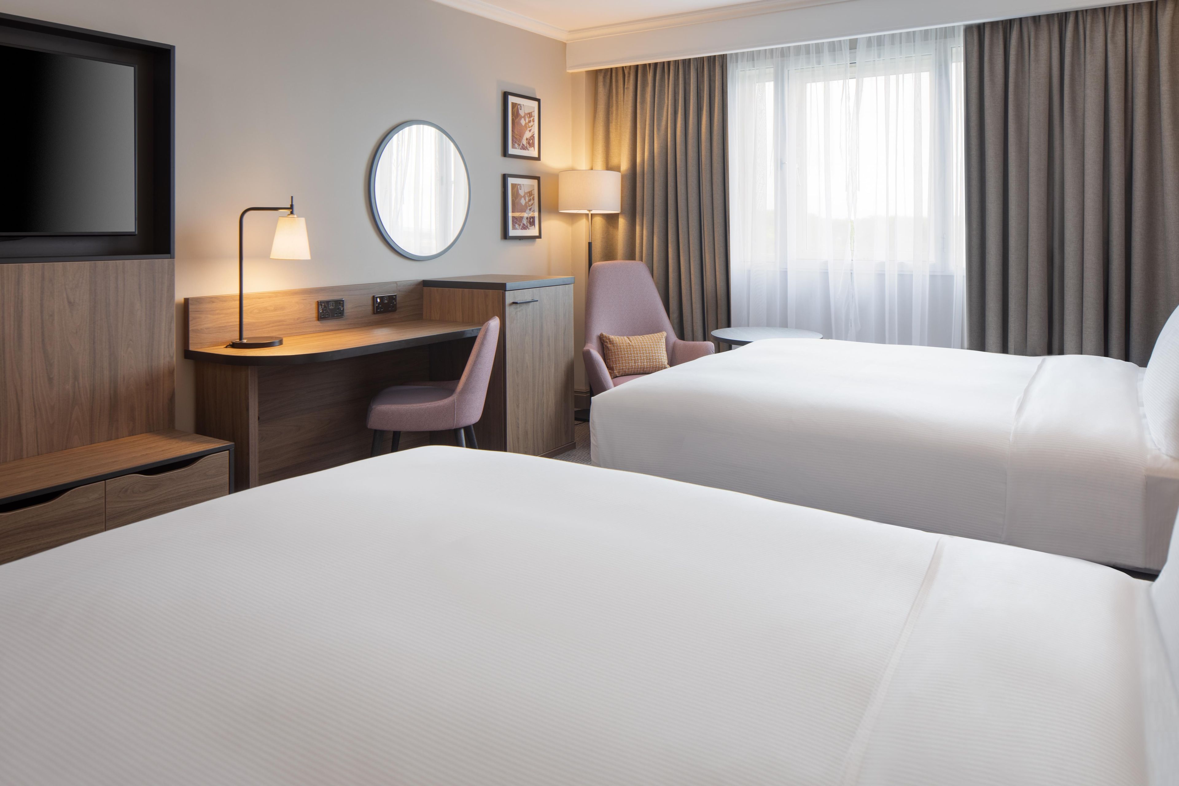 Habitación de hotel con dos camas, un escritorio, una silla, un televisor y un ventanal con cortinas.