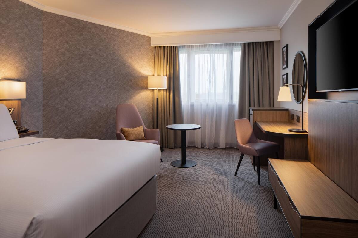 Een hotelkamer met een groot bed, een bureau, een stoel, een klein tafeltje, een televisie en een lamp.