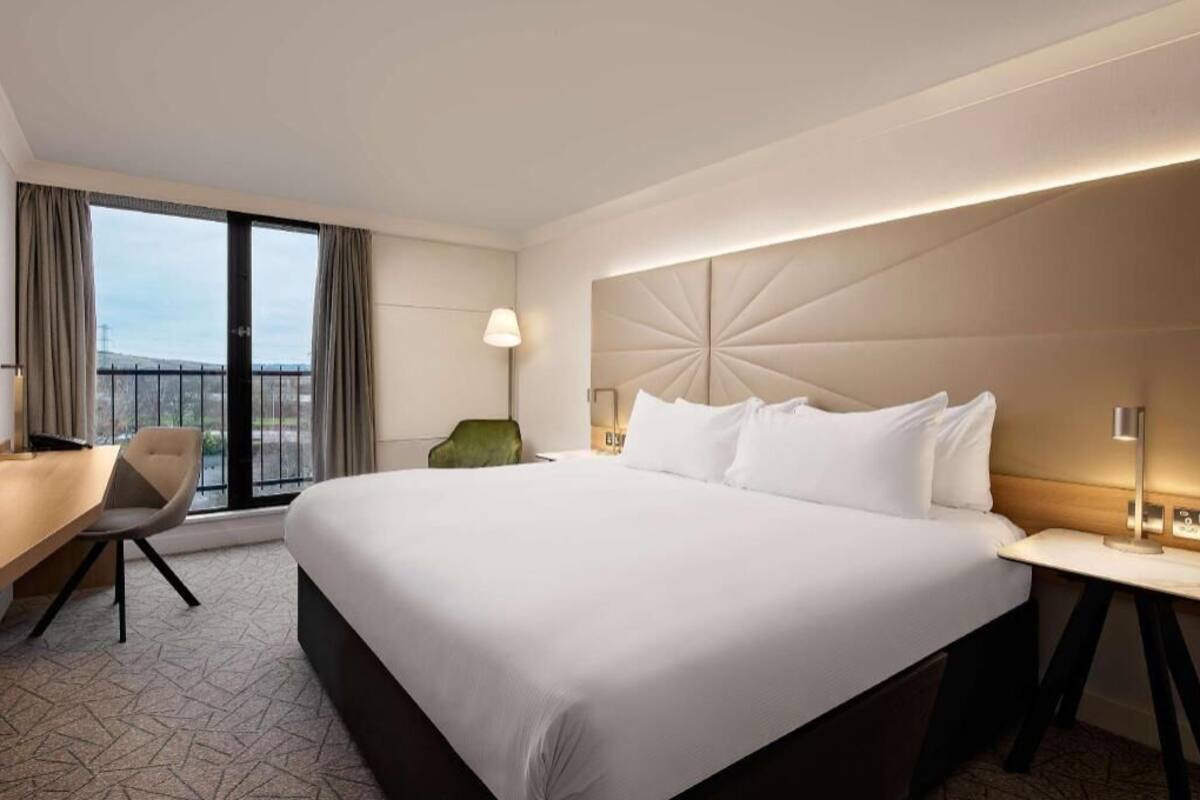 A hotel room with a large bed, a desk, a chair, a lamp, and a view of the outdoors.