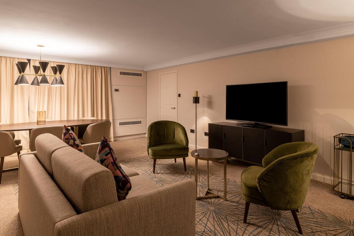 A modern living room with a sofa, two armchairs, a small round table, a flat-screen TV, and a dining area with a table and chairs.