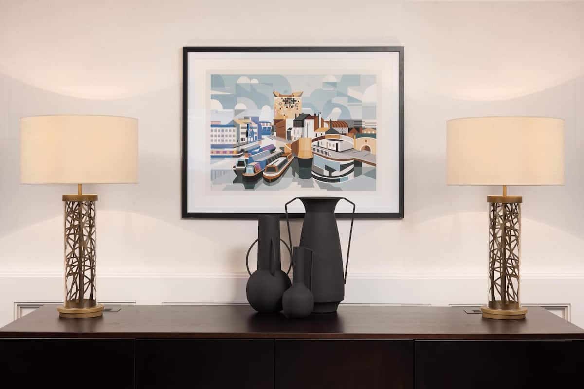 A framed cityscape artwork, two modern lamps, and a vase on a dark wooden table.