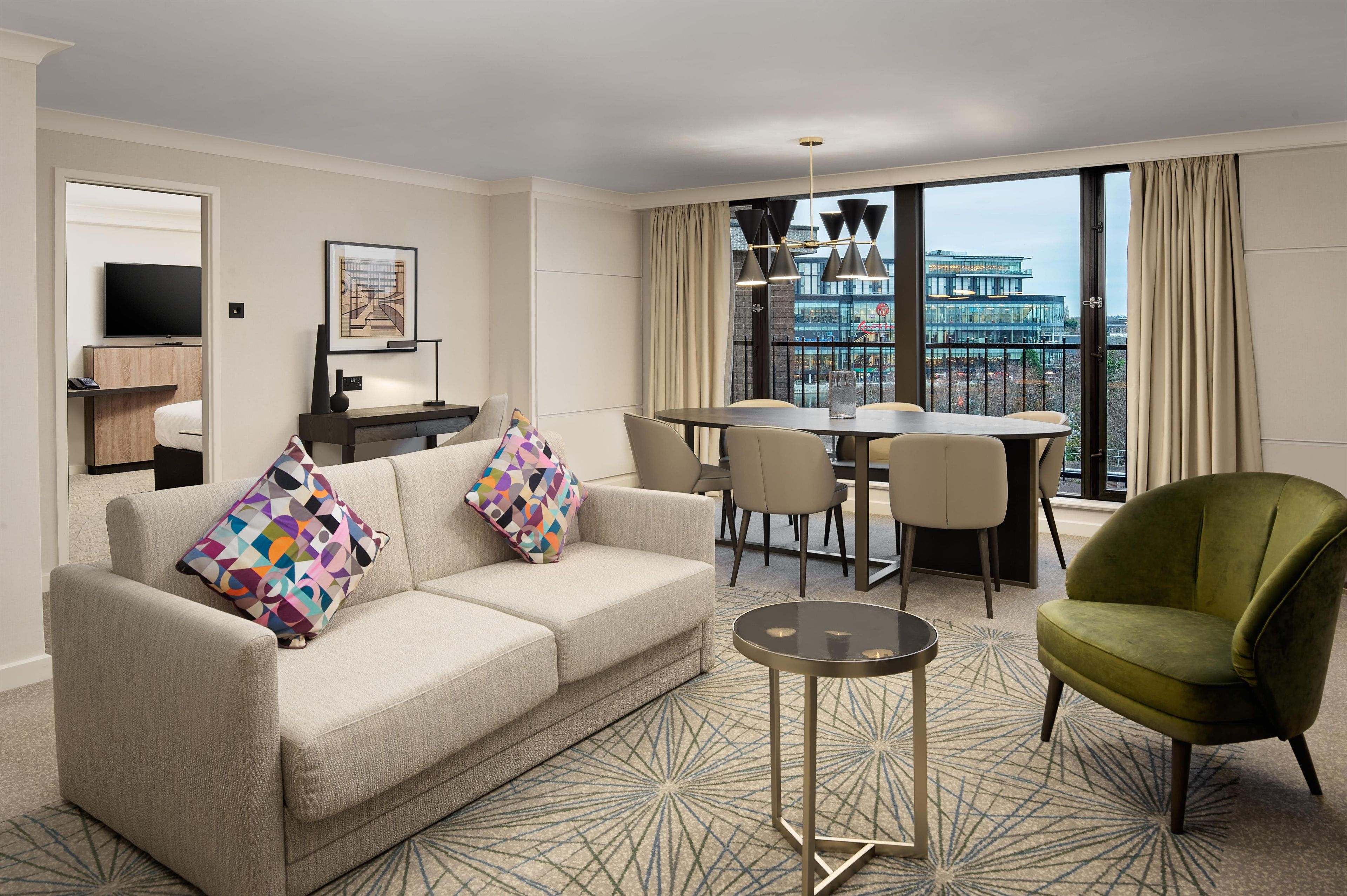 Habitación de hotel con sofá, un sillón verde, una mesa de comedor con sillas, un televisor y vistas a la ciudad a través de amplias ventanas.