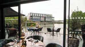 Un área de comedor al aire libre con mesas y sillas, vista a un edificio moderno a través de un cuerpo de agua, y una planta en maceta a la derecha.