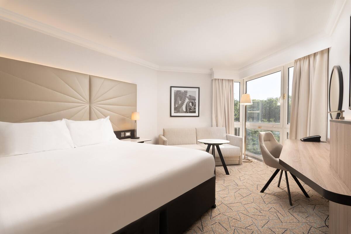A hotel room with a large bed, a desk, a chair, a small table, a lamp, a framed picture, and a view of the outdoors.