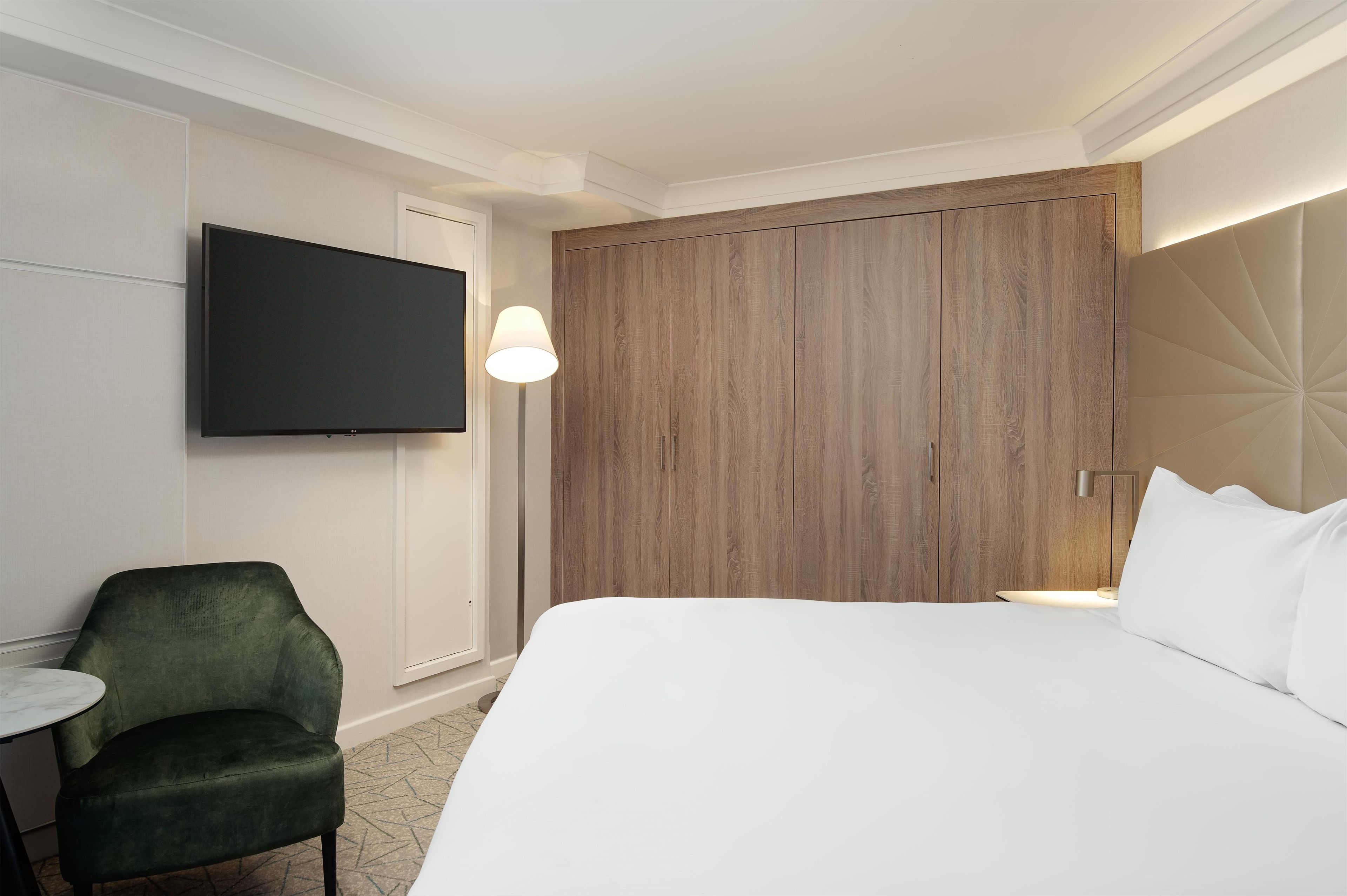 Habitación de hotel con cama, televisor montado en la pared, escritorio, silla y armario de madera.
