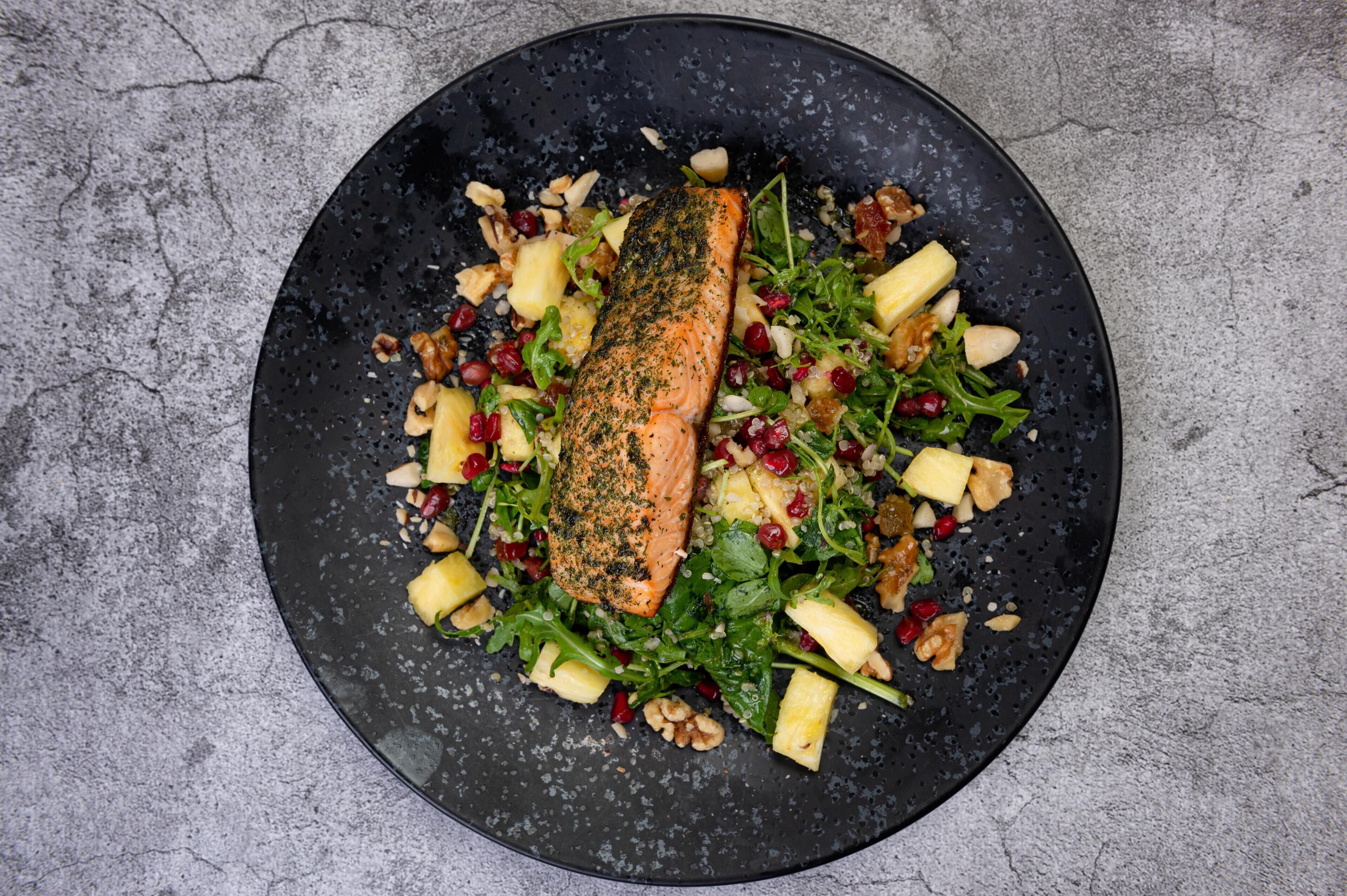 Ein schwarzer Teller mit Lachsfilet, Ananasstückchen, Walnüssen und grünem Salat.