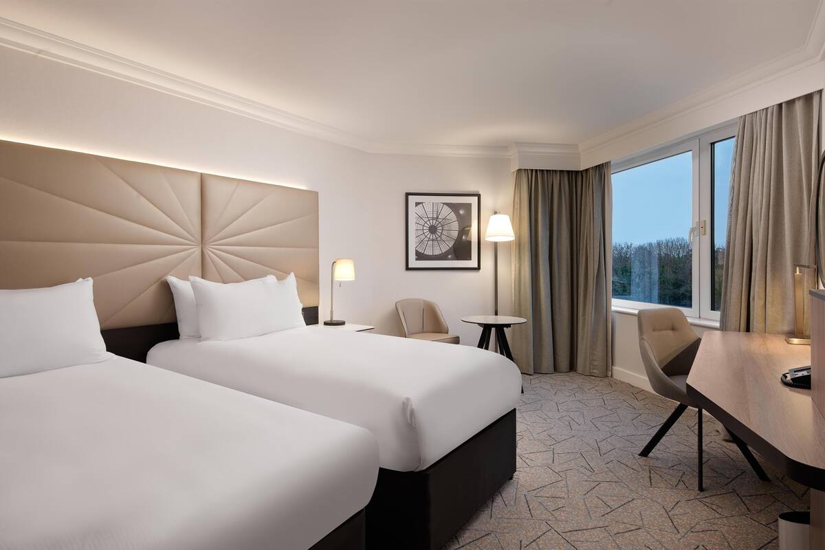 A hotel room with two beds, a desk, a chair, a lamp, and a window with curtains.