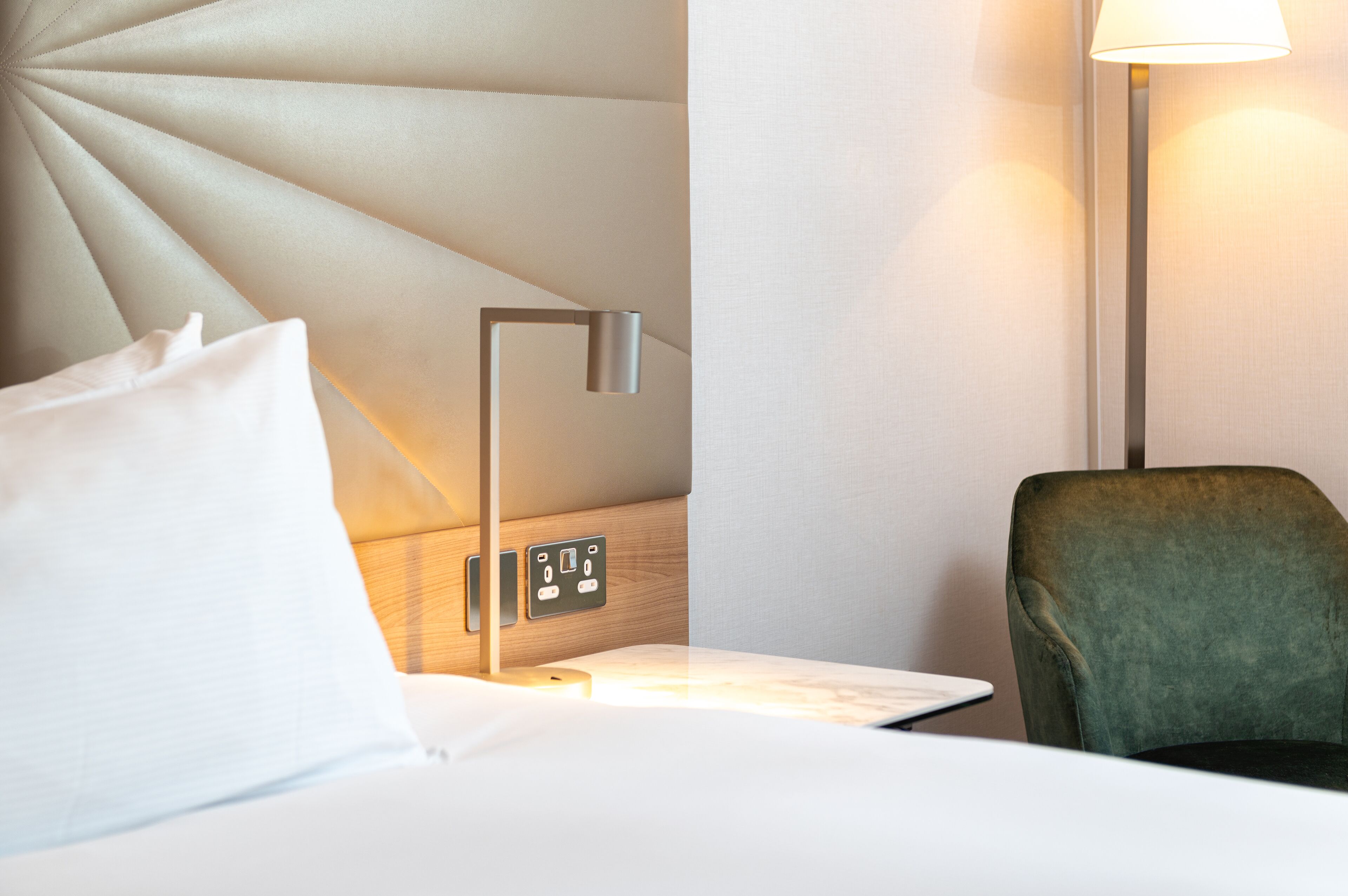 Ein Hotelzimmer mit Bett, einer Nachttischlampe, einem grünen Sessel und einer Wandbedienung.