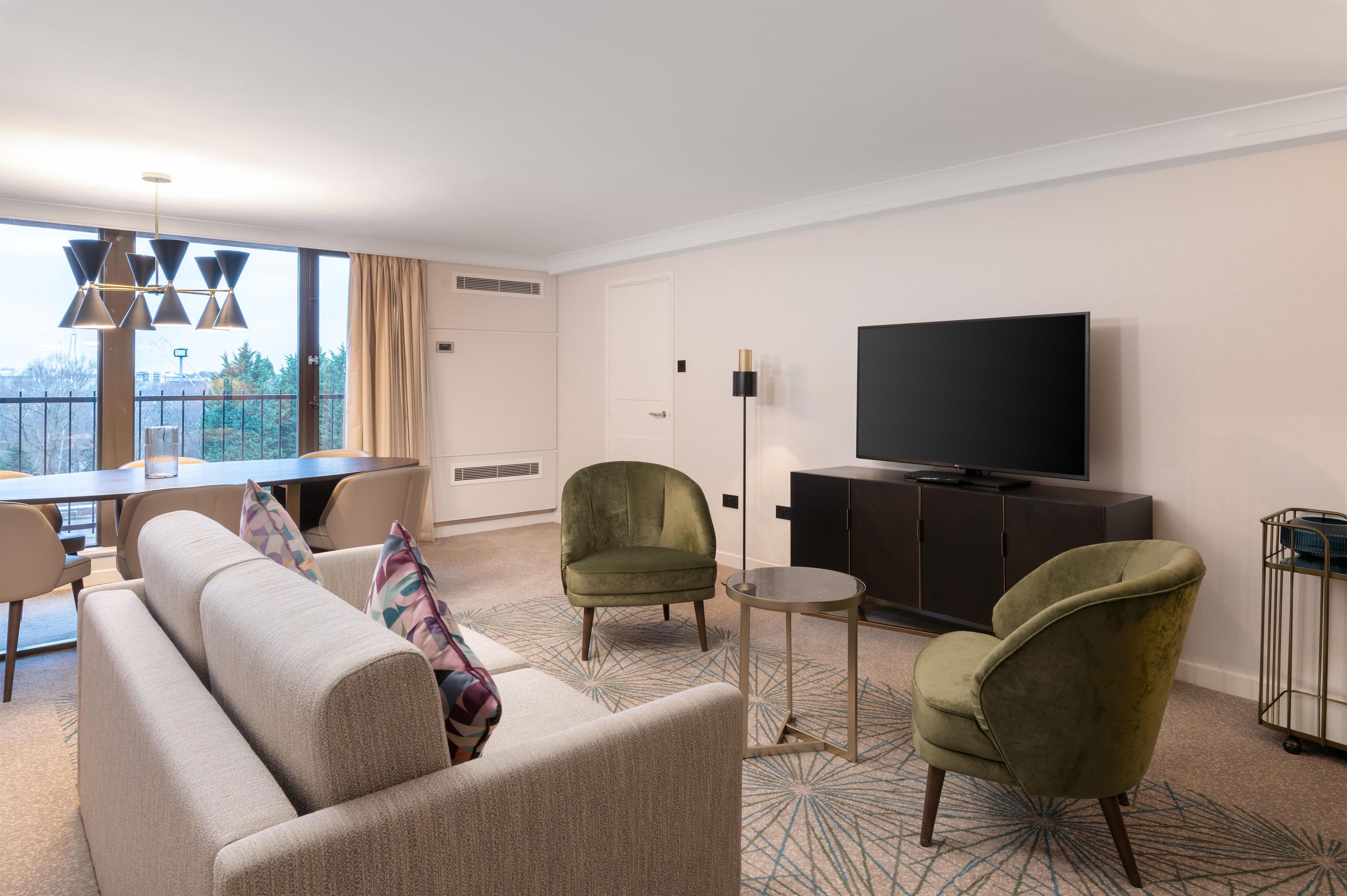 A hotel room with a sofa, two armchairs, a dining table, a TV, and a balcony with a view.
