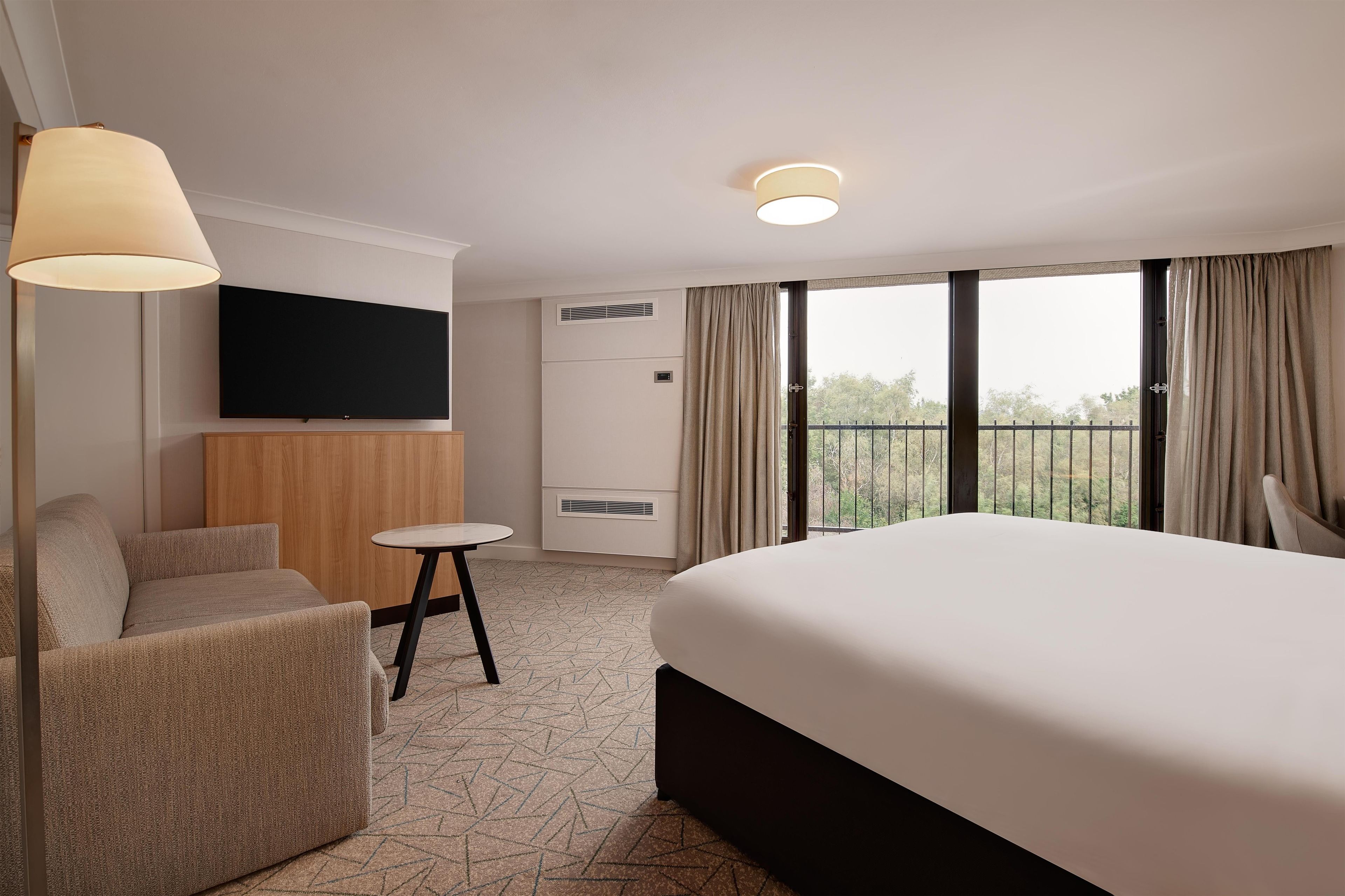 A hotel room with a bed, a TV, a small table, a sofa, and a balcony with curtains.