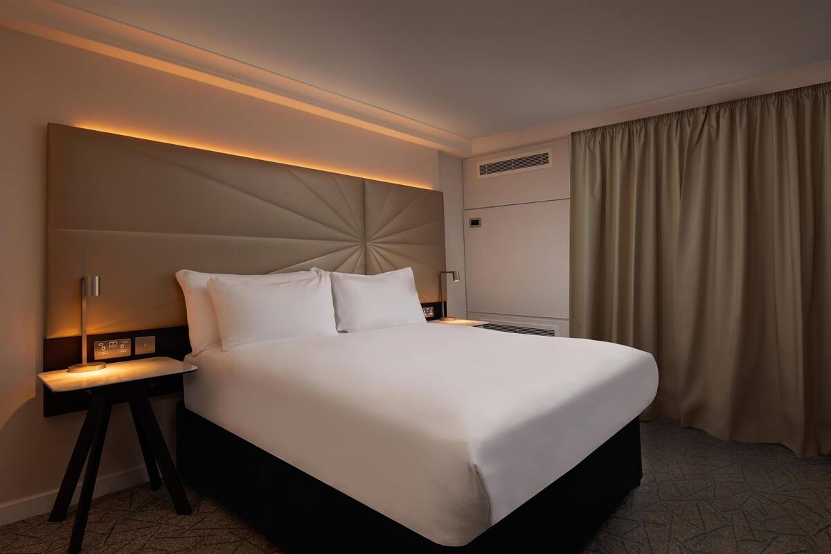 A hotel room with a large bed, a bedside table with a lamp, a headboard with a light panel, and a window with curtains.